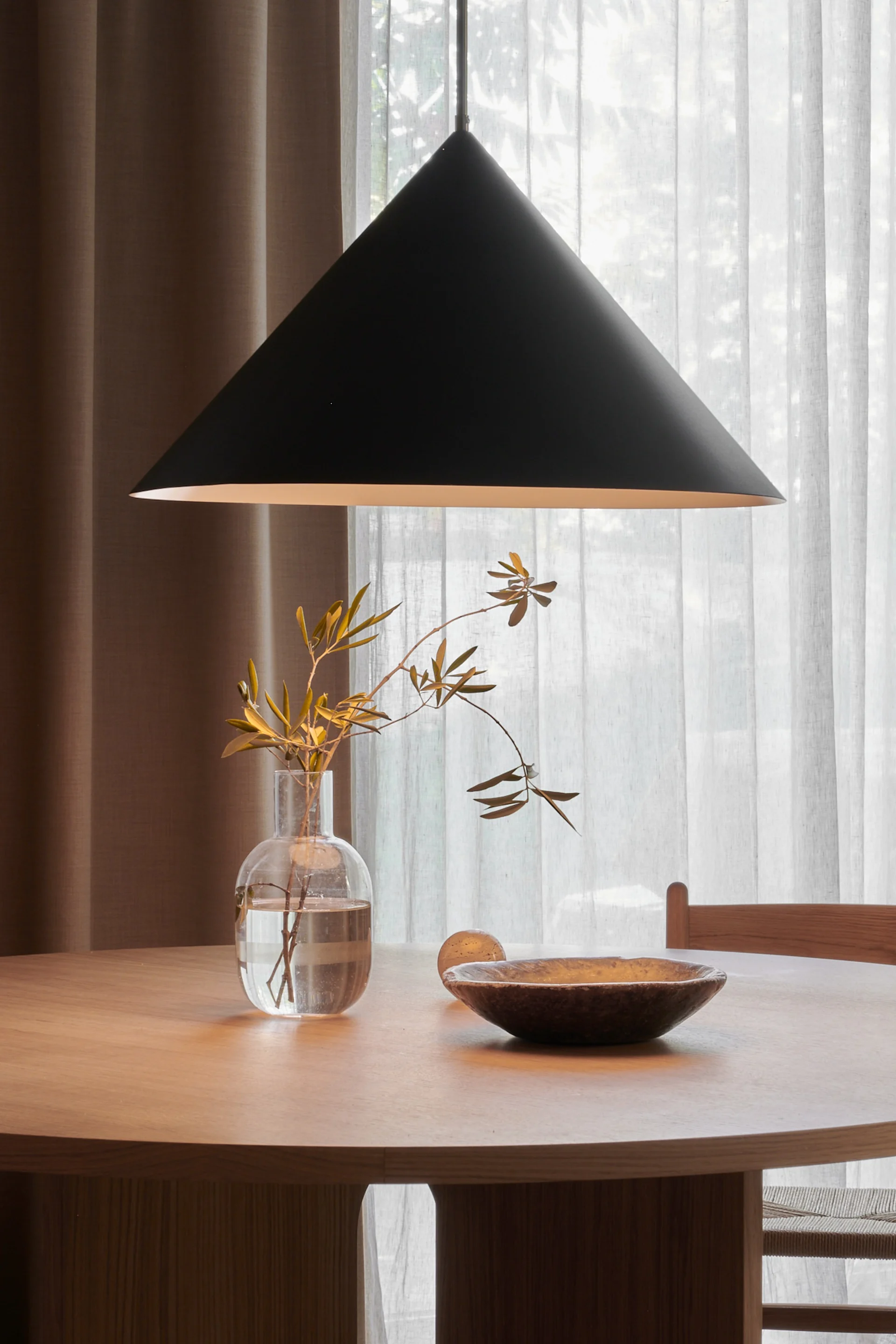 Runder Holzesstisch mit schwarzer konischer Pendelleuchte darüber, mit einer klaren Vase mit Ästen und einer dunklen Holzschale.