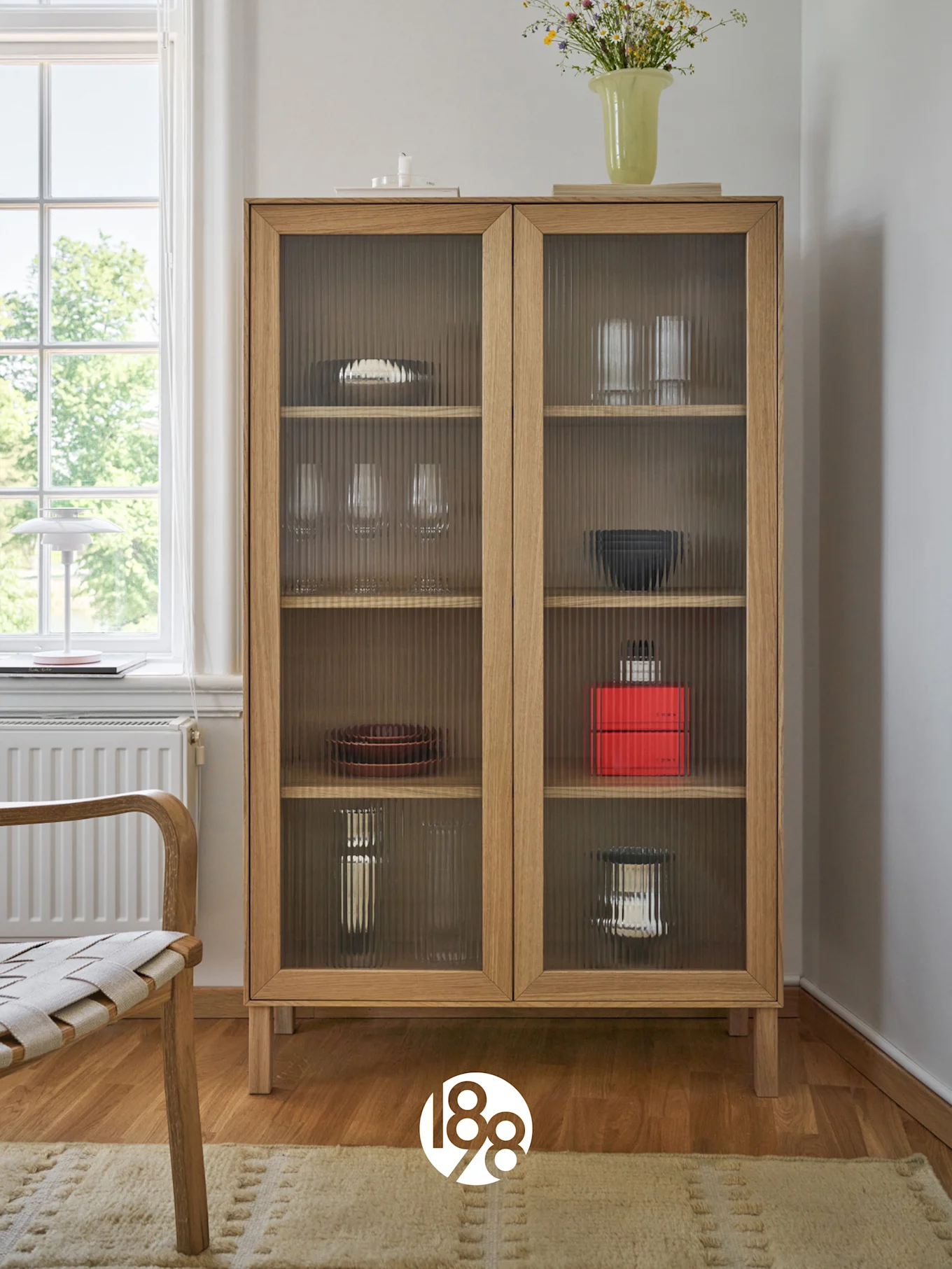 Ein heller Holzschrank mit geriffelten Glastüren, in dem Geschirr und Gläser stehen, darauf eine Vase mit Wildblumen. Ein Stuhl und ein Fenster sind links.