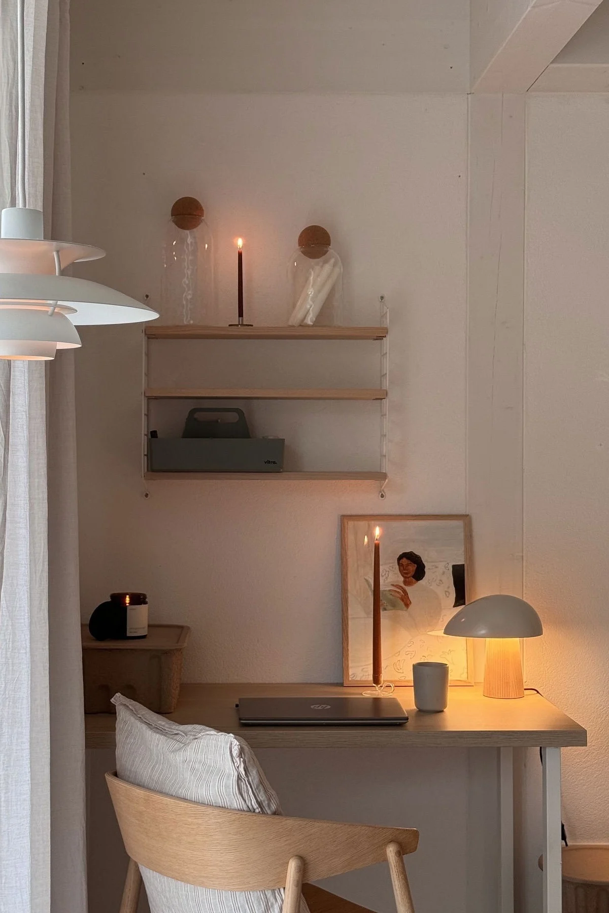 Ein gemütlicher Schreibtisch mit warmer Beleuchtung, einem geschlossenen Laptop, zwei brennenden Kerzen, einer modernen Lampe und einem gerahmten Kunstwerk.