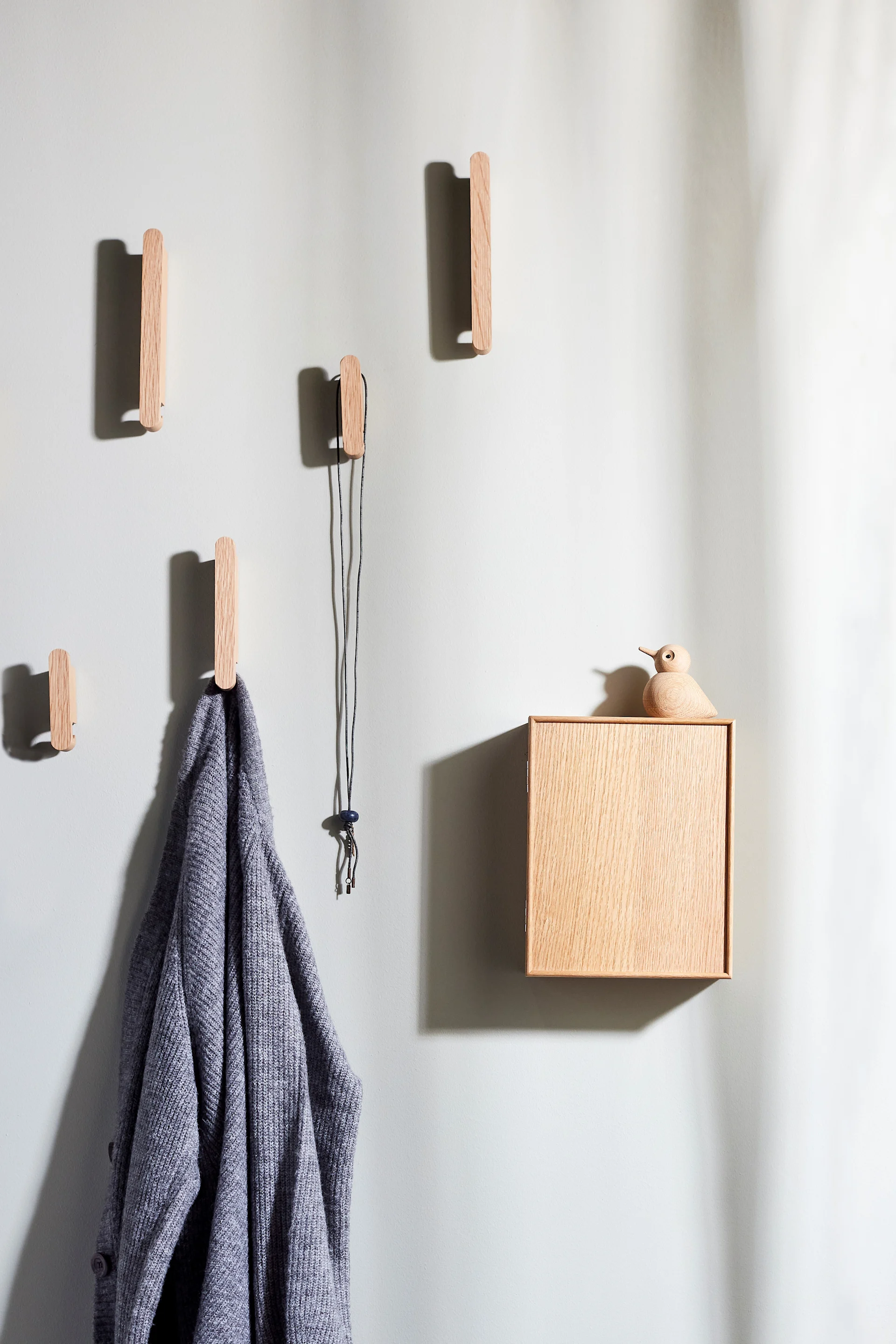 Haken aus Holz, ein grauer Cardigan und eine Holzkiste mit einer Vogelfigur an einer hellen Wand.