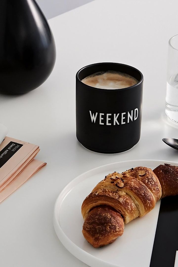 Kaffeebecher: Der schwarze Kaffeebecher mit dem Aufdruck "Weekend" von Design Letters steht auf einem Tisch, davor ein Teller mit einem Croissant.