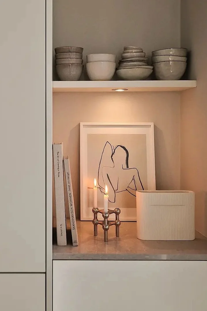 Ein gemütliches Regal mit Keramikschalen, Büchern, gerahmter Kunst, angezündeten Kerzen in einem Messinghalter und einer geriffelten Vase.