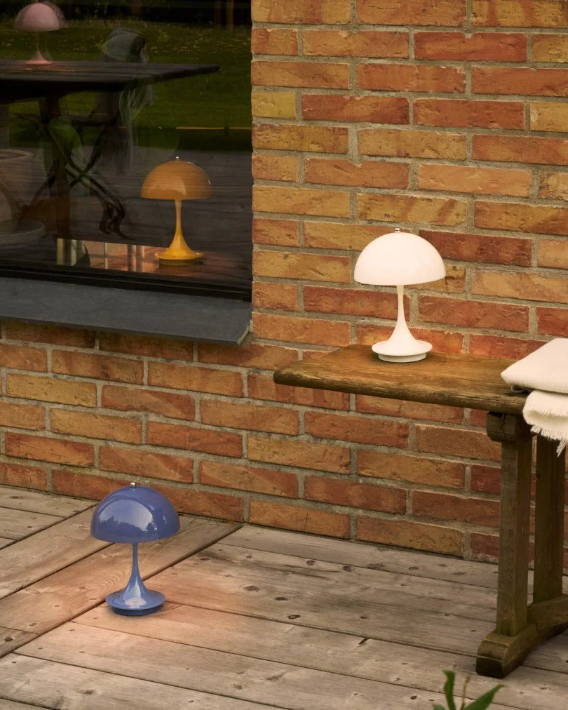 Drei pilzförmige Lampen in Weiß, Blau und Gelb auf einer Holzterrasse an einer Ziegelwand und Fenster.