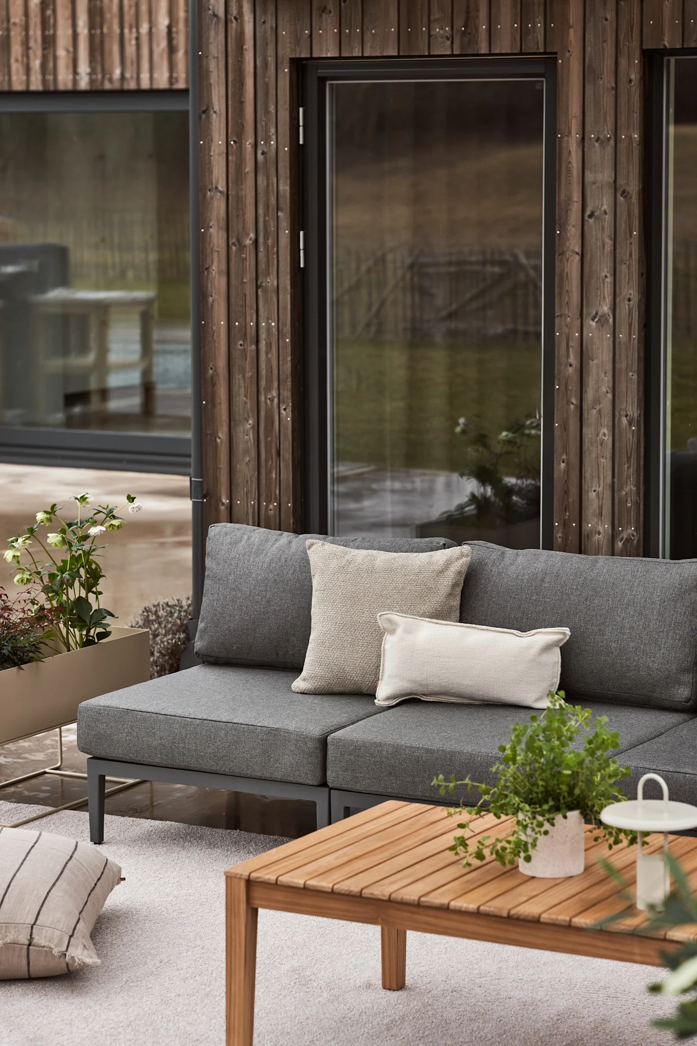 Ein Außenwohnbereich mit einem grauen Modulsofa, hellen Kissen, einem Holztisch, einem hellen Teppich und Pflanzgefäßen mit Grün vor einem modernen Holzgebäude.