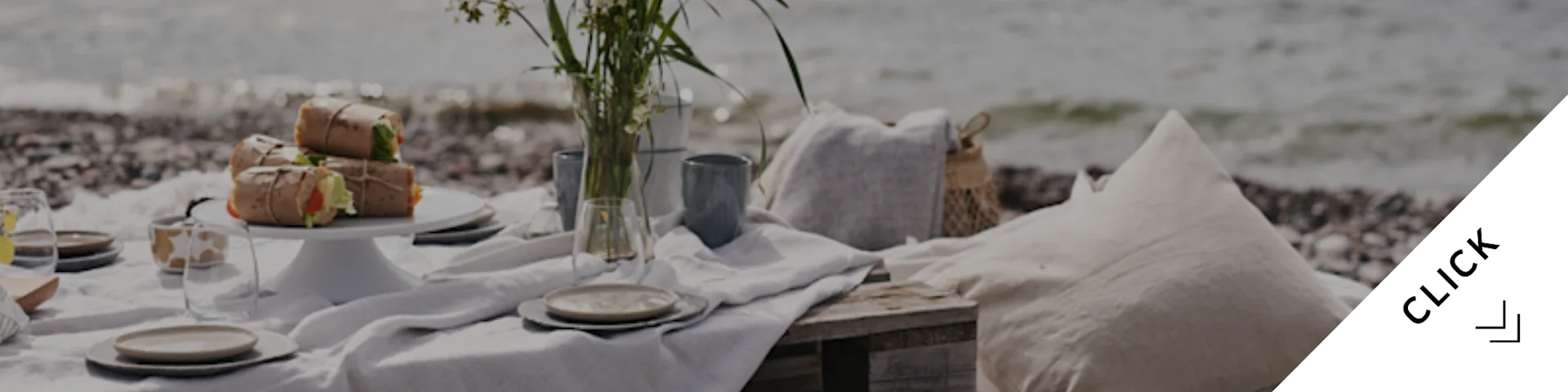 Ein idyllisches Strandpicknick mit eingepacktem Essen, Geschirr, einer Blumenvase und Kissen an einem Kieselstrand am Meer.