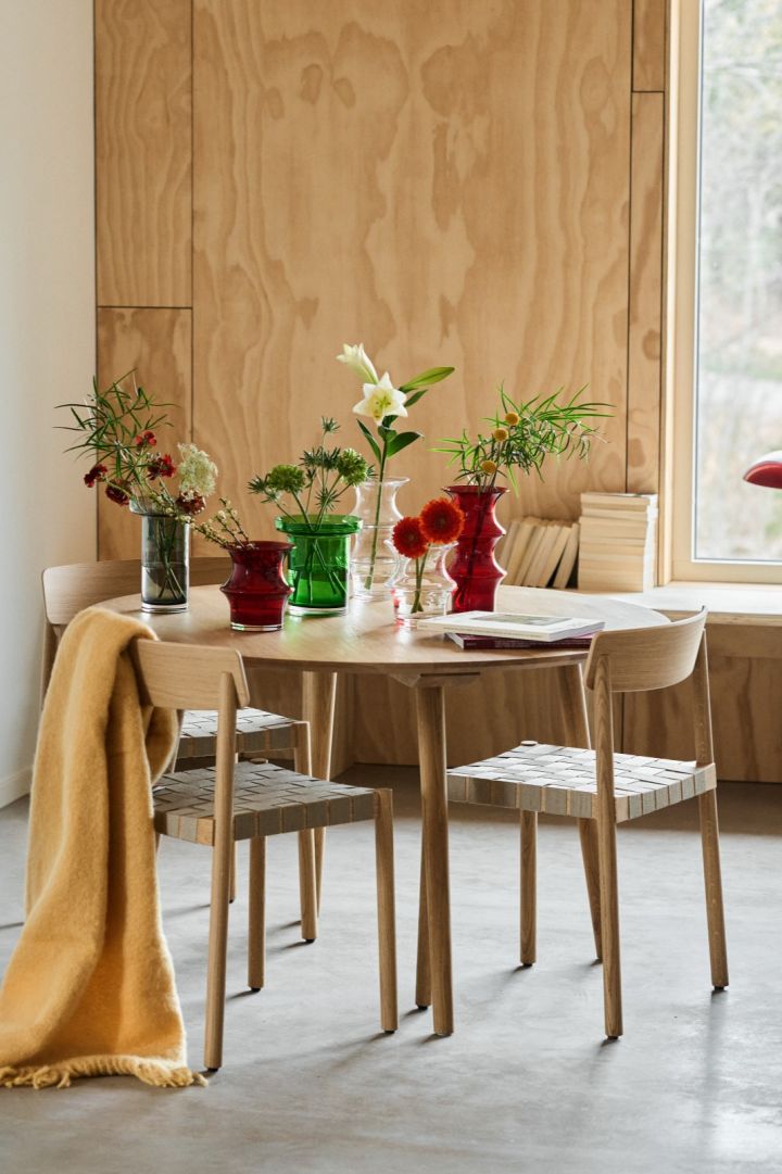 Ein gedeckter Tisch steht inmitten eines mit Holz ausgekleideten Raums. Darauf befinden sich mehrere Vasen in Rot, Grün, Grau und Durchsichtig.