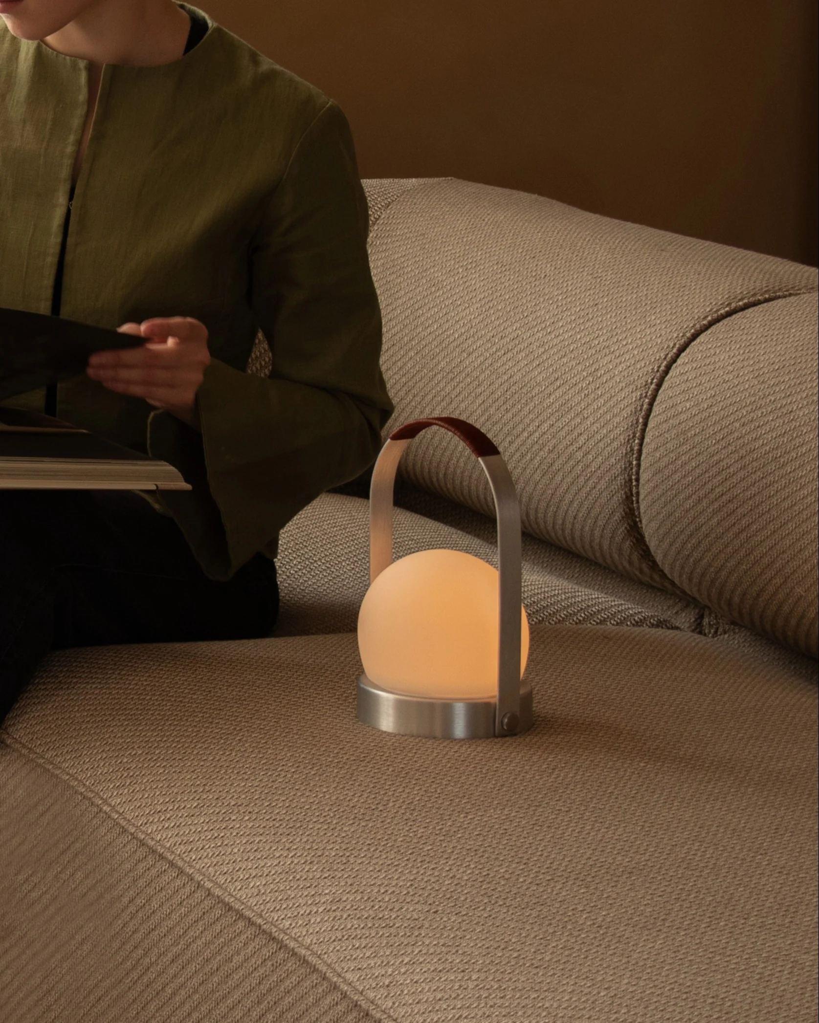 Eine Person liest ein Buch auf einem beigen Sofa neben einer leuchtenden kugelförmigen tragbaren Lampe mit Ledergriff.