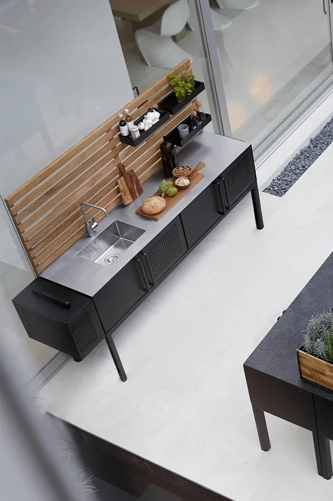 Moderne Außenküche mit Edelstahlspüle, schwarzen Schränken, Holzwandverkleidung und Regalen mit Gewürzen und Lebensmitteln.