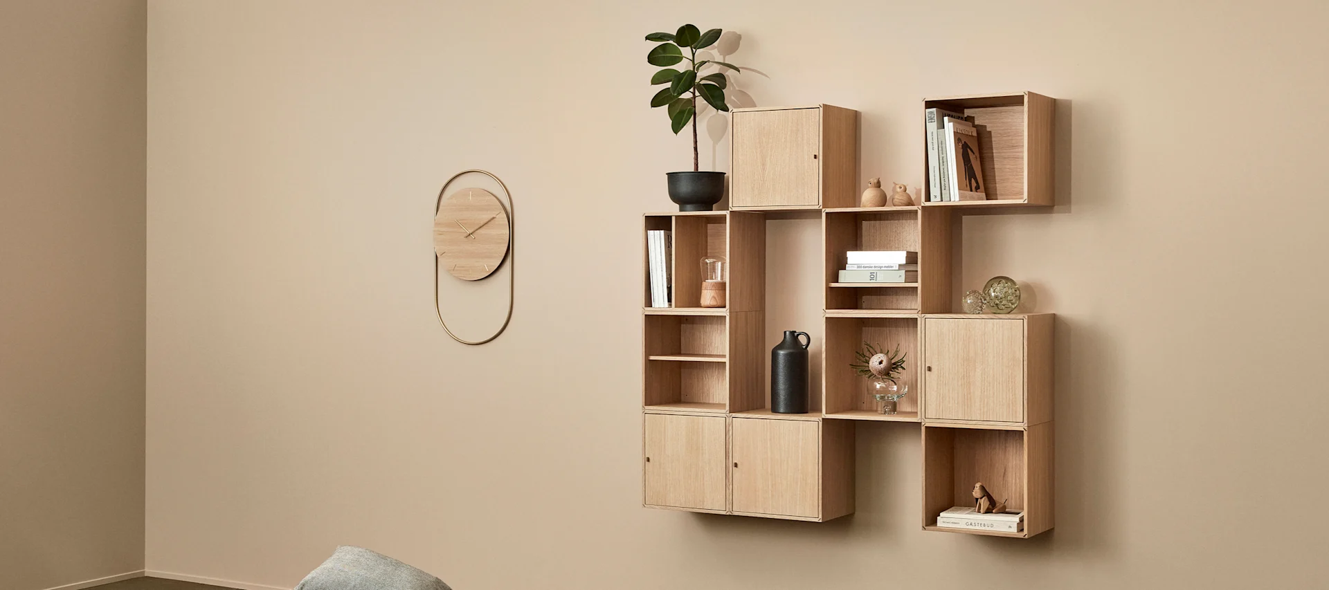 Modulare Holzregaleinheit mit Dekoartikeln und einer goldenen ovalen Wanduhr an einer hellbraunen Wand.
