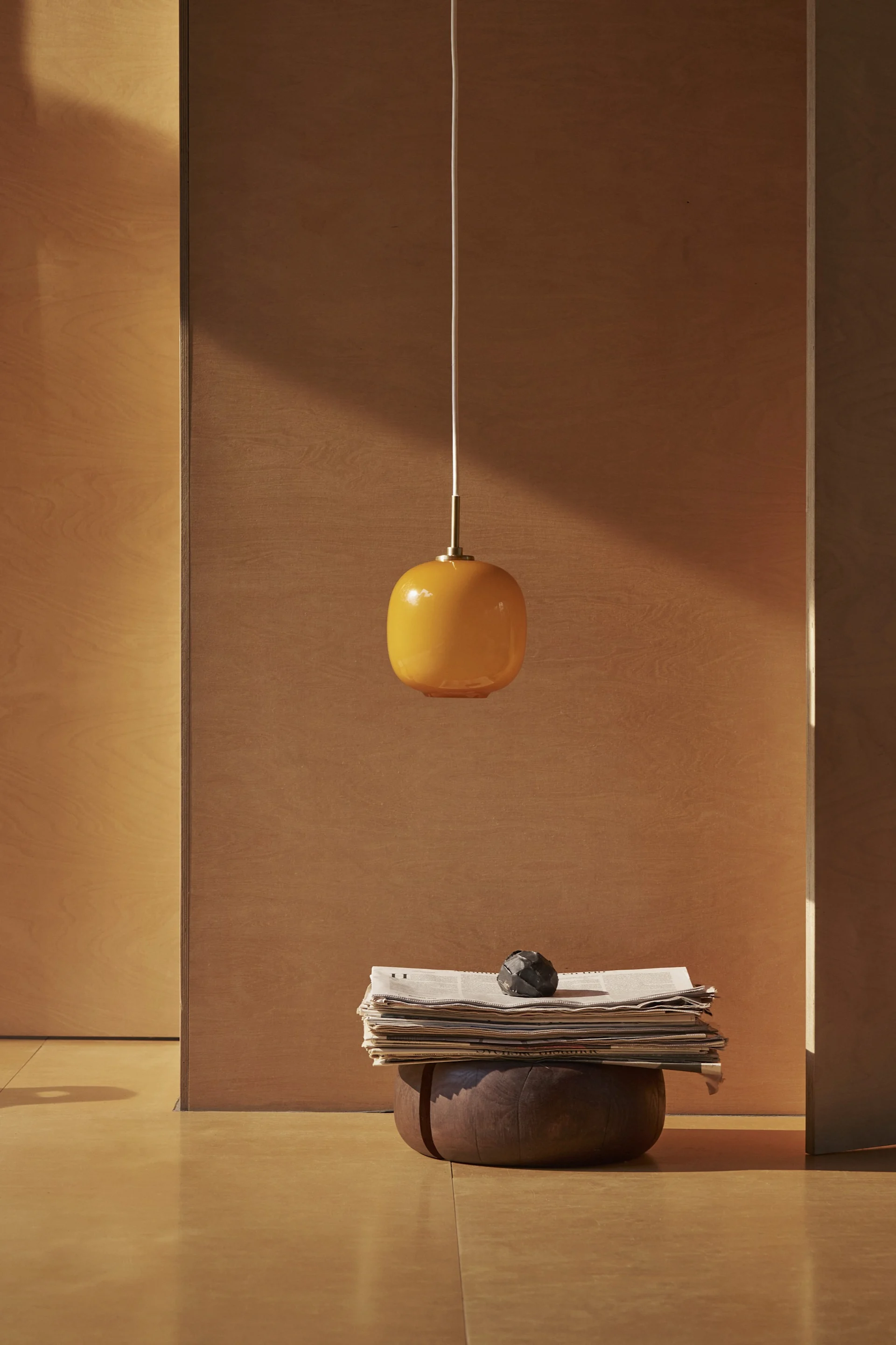 Eine orangefarbene ovale Pendelleuchte hängt über einem Stapel Zeitungen und einem dunklen Deko-Objekt auf einem Holzsockel in einem minimalistischen Raum.