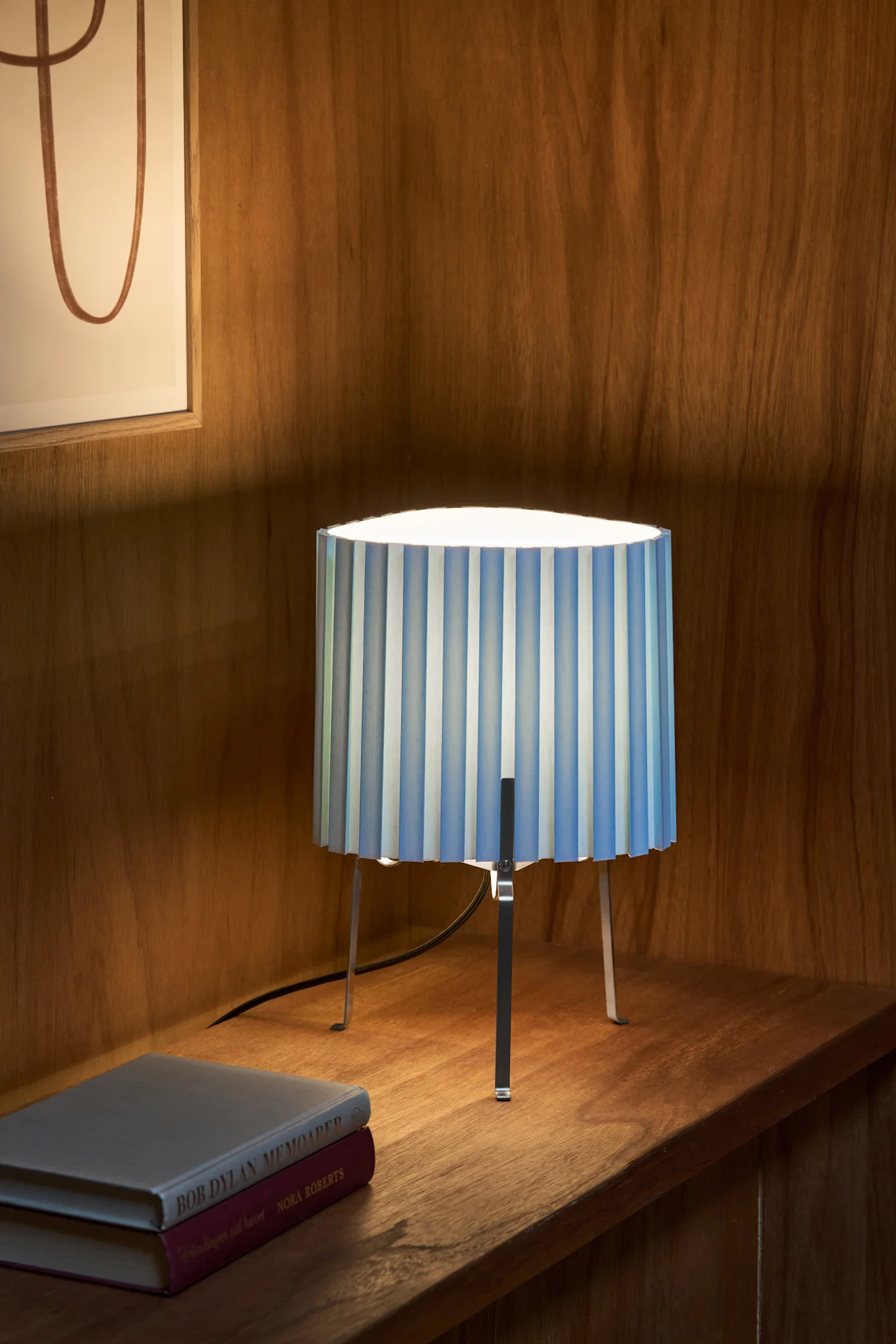 Eine Tischlampe mit einem blau-weiß gestreiften plissierten Schirm und Metallbeinen, die auf einem Holzregal neben zwei Büchern steht.