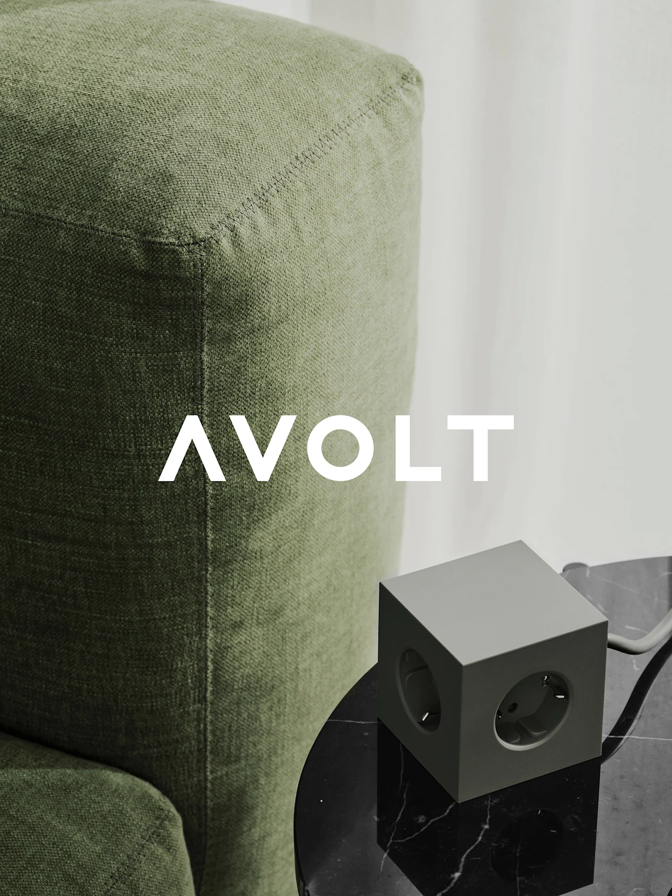 Eine grüne würfelförmige Steckdosenleiste mit drei Ausgängen auf einem schwarzen Marmortisch neben einem grünen Sofa. AVOLT-Logo.