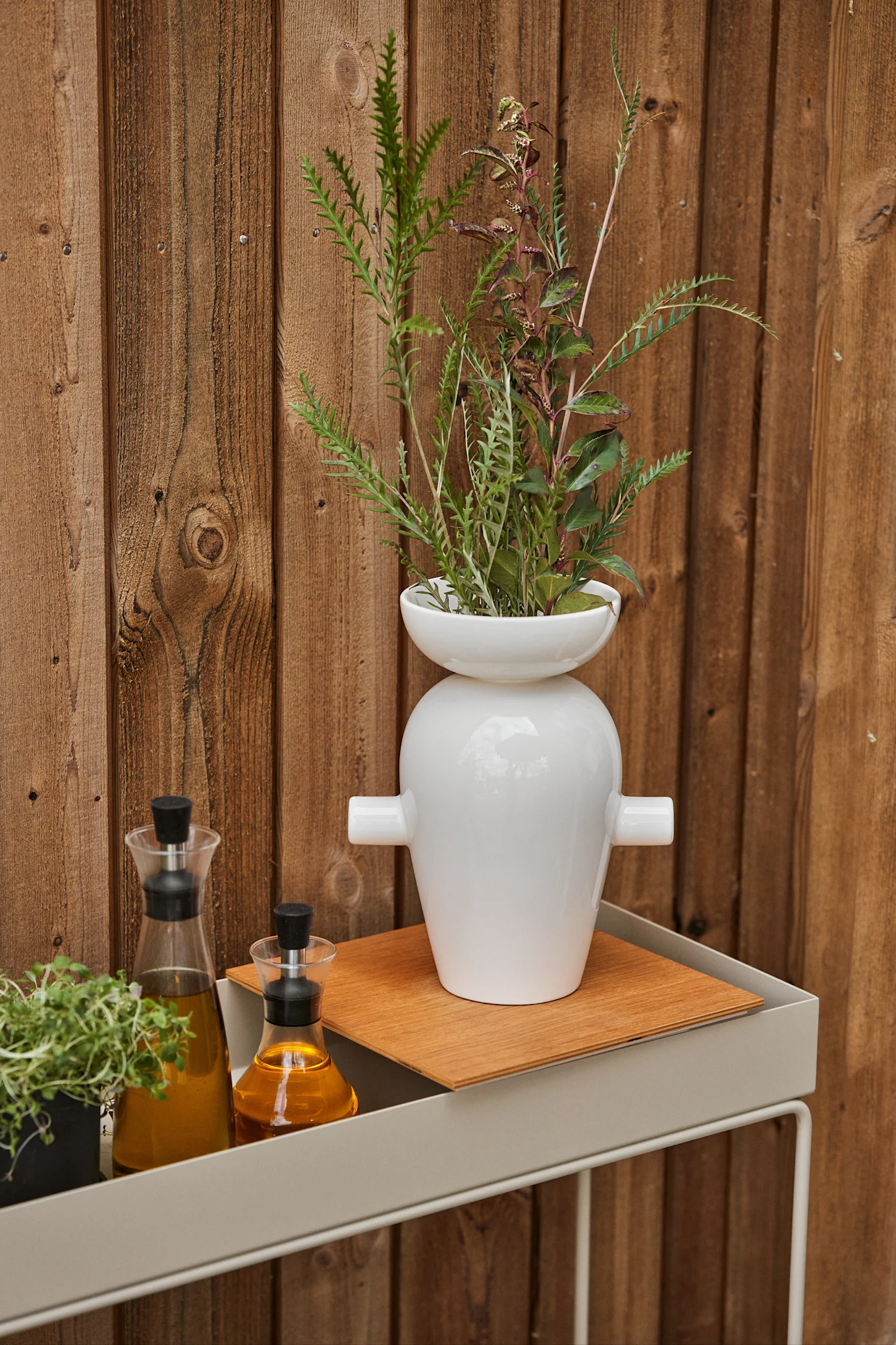 Weiße Vase mit Laub, zwei Ölflaschen und einem Kräutertopf auf einem grauen Regal vor einem Holzzaun.