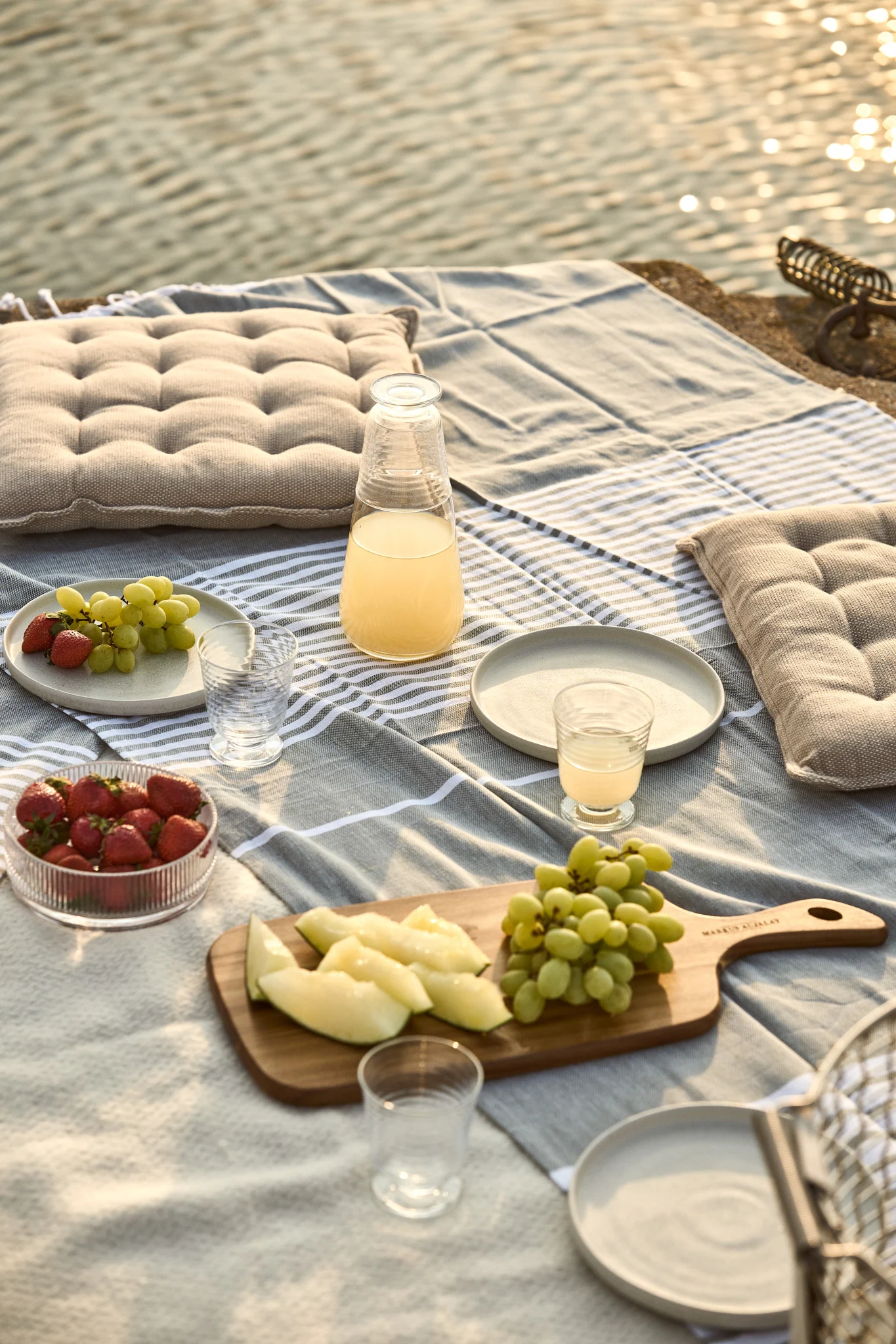 Picknick am Wasser mit gestreifter Decke, Kissen, Saft, Erdbeeren, Trauben und Melonenscheiben.