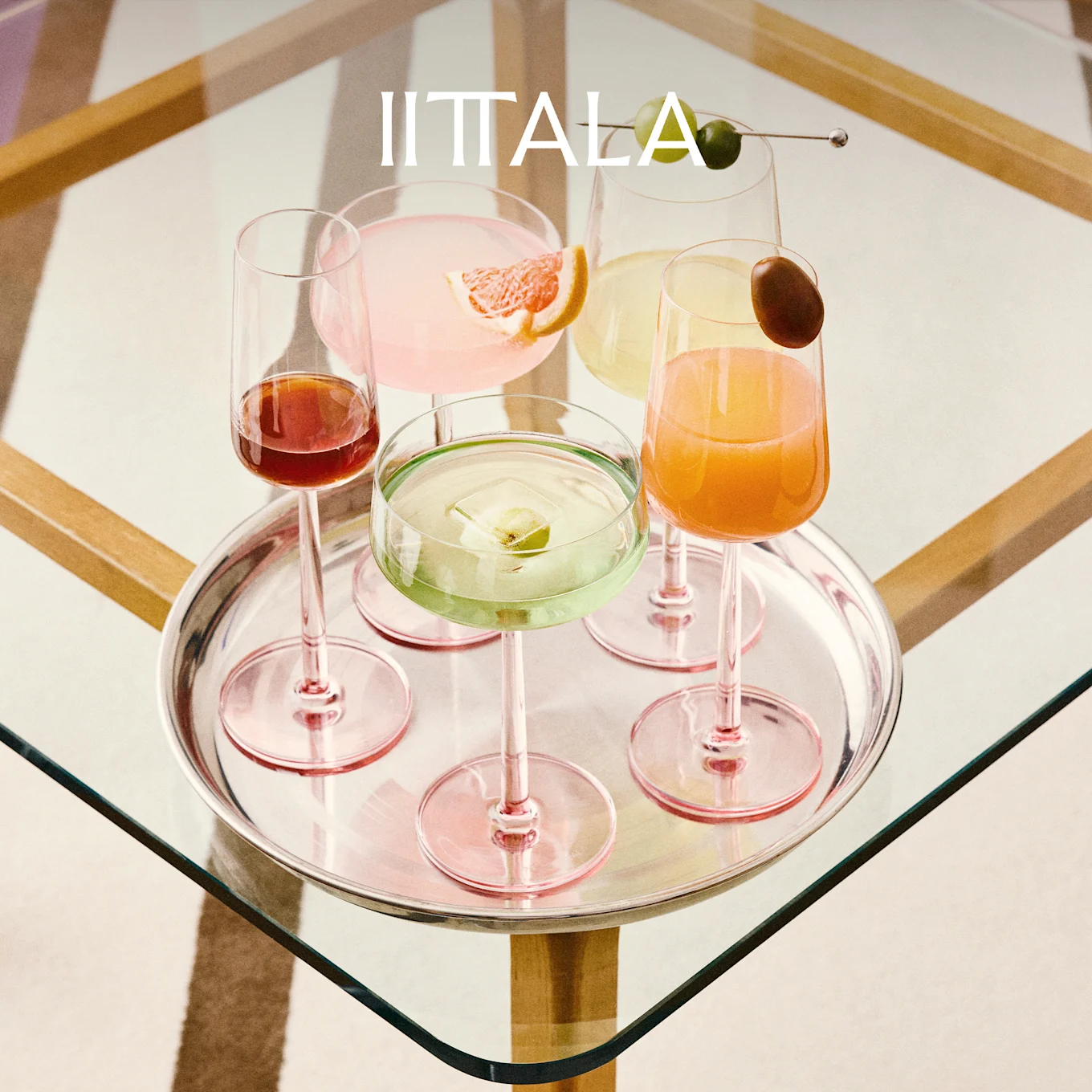 IITTALA-Logo über einem Silbertablett mit fünf farbenfrohen Cocktails in rosa gestielten Gläsern auf einem Glastisch.