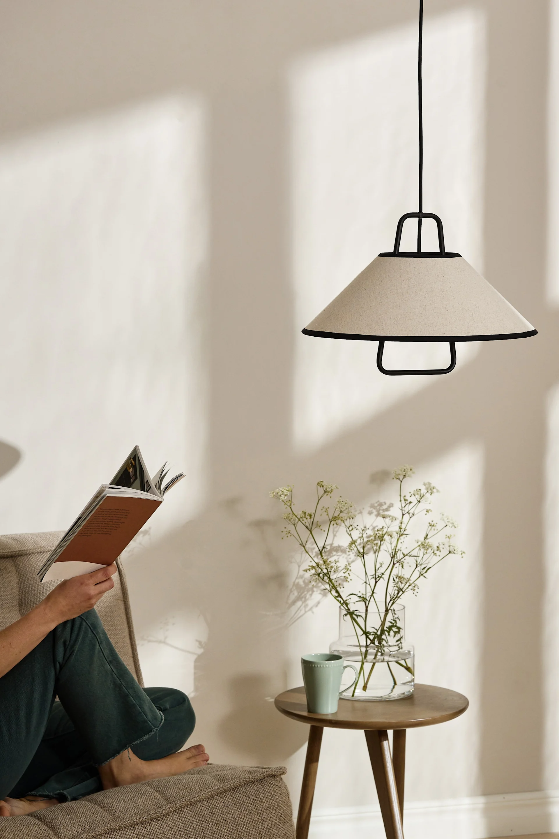 Eine Person liest ein Buch in einem Sessel neben einem runden Holztisch mit einer Glasvase mit Blumen, einer Tasse und einer beigefarbenen Pendelleuchte.