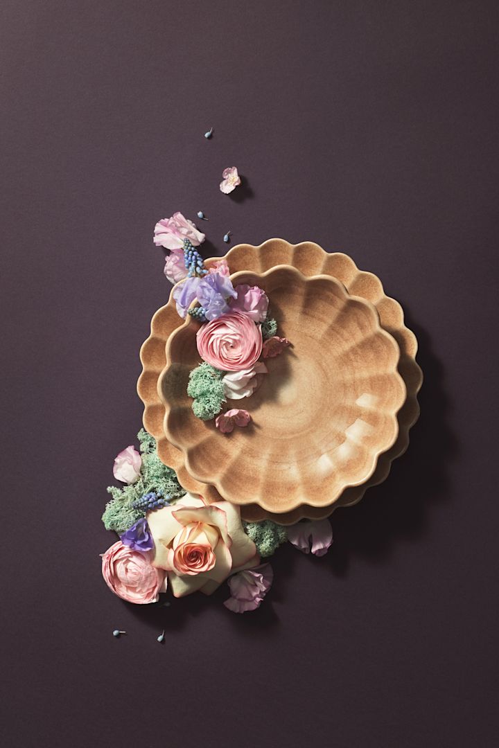 Vor einem braunen Hintergrund wurden ein Teller und eine Schale aus der Oyster-Kollektion in der Farbe Cinnamon von Mateus mit Blumen kunstvoll in Szene gesetzt.