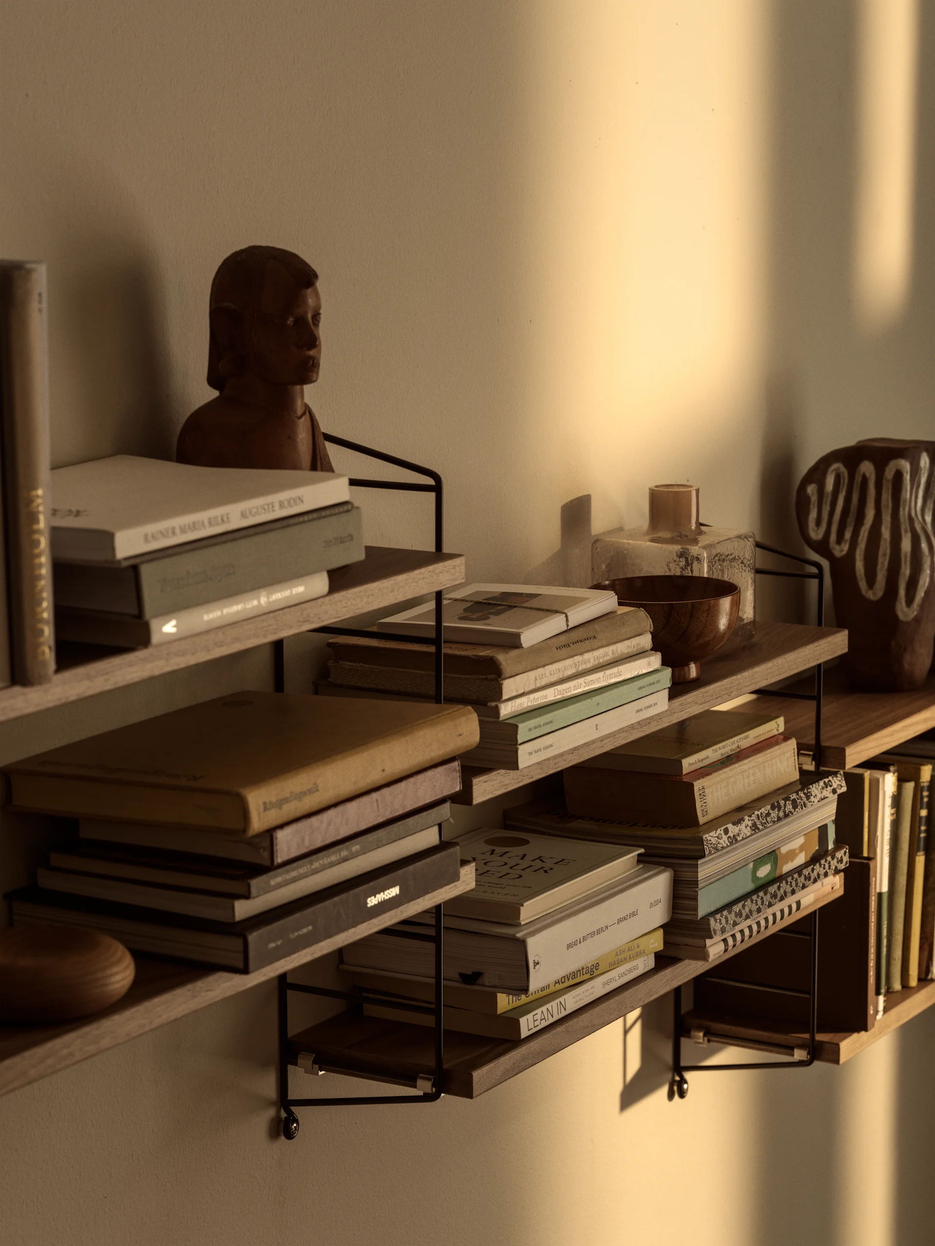 Wandmontierte Regale gefüllt mit Büchern, einer Holzbüste und Dekorationsgegenständen, beleuchtet von warmem Sonnenlicht an der Wand.