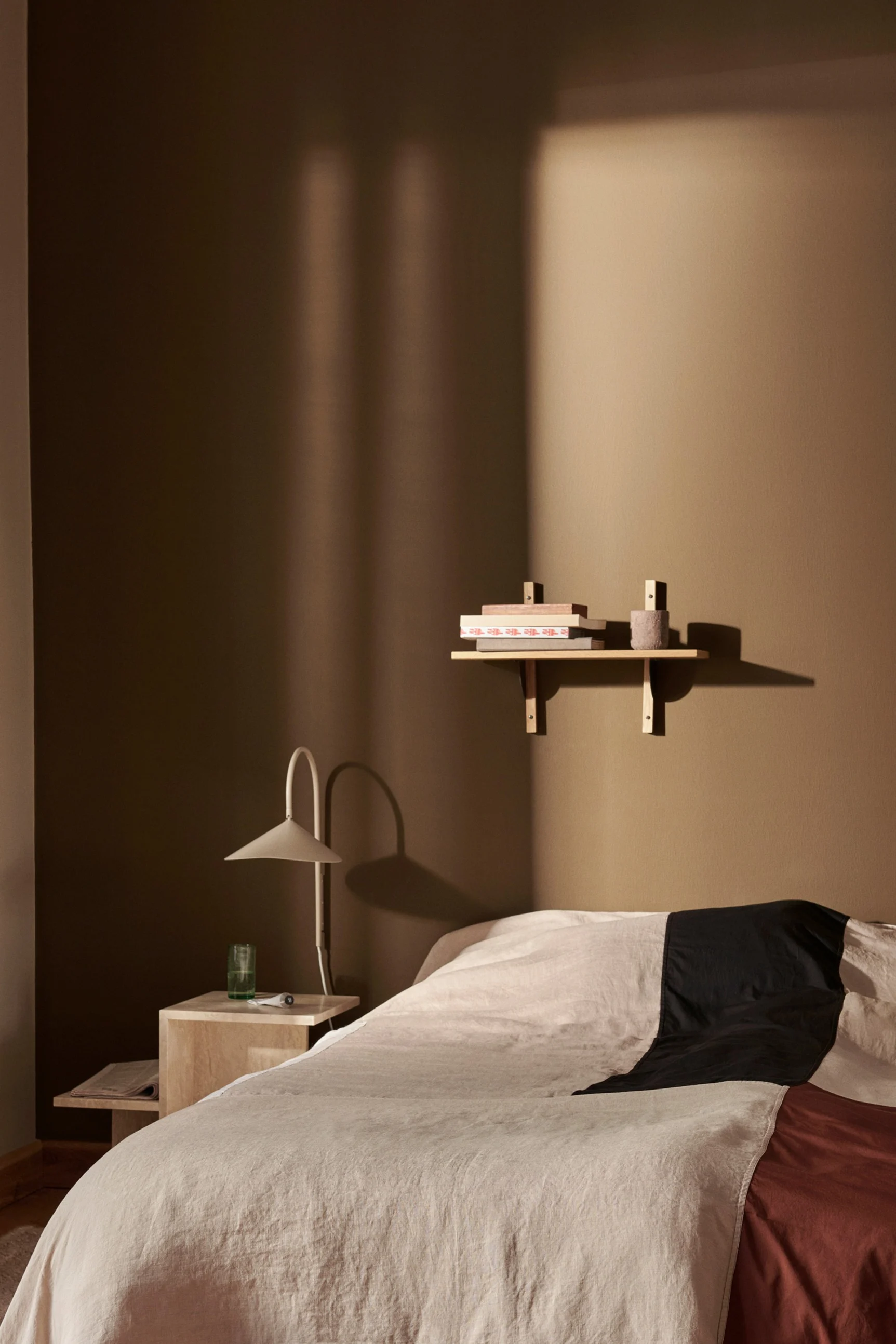 Ein Schlafzimmer mit einem Bett mit beiger, schwarzer und rostfarbener Bettwäsche, einem Nachttisch mit Lampe und einem Regal mit Büchern an einer warmbraunen Wand.