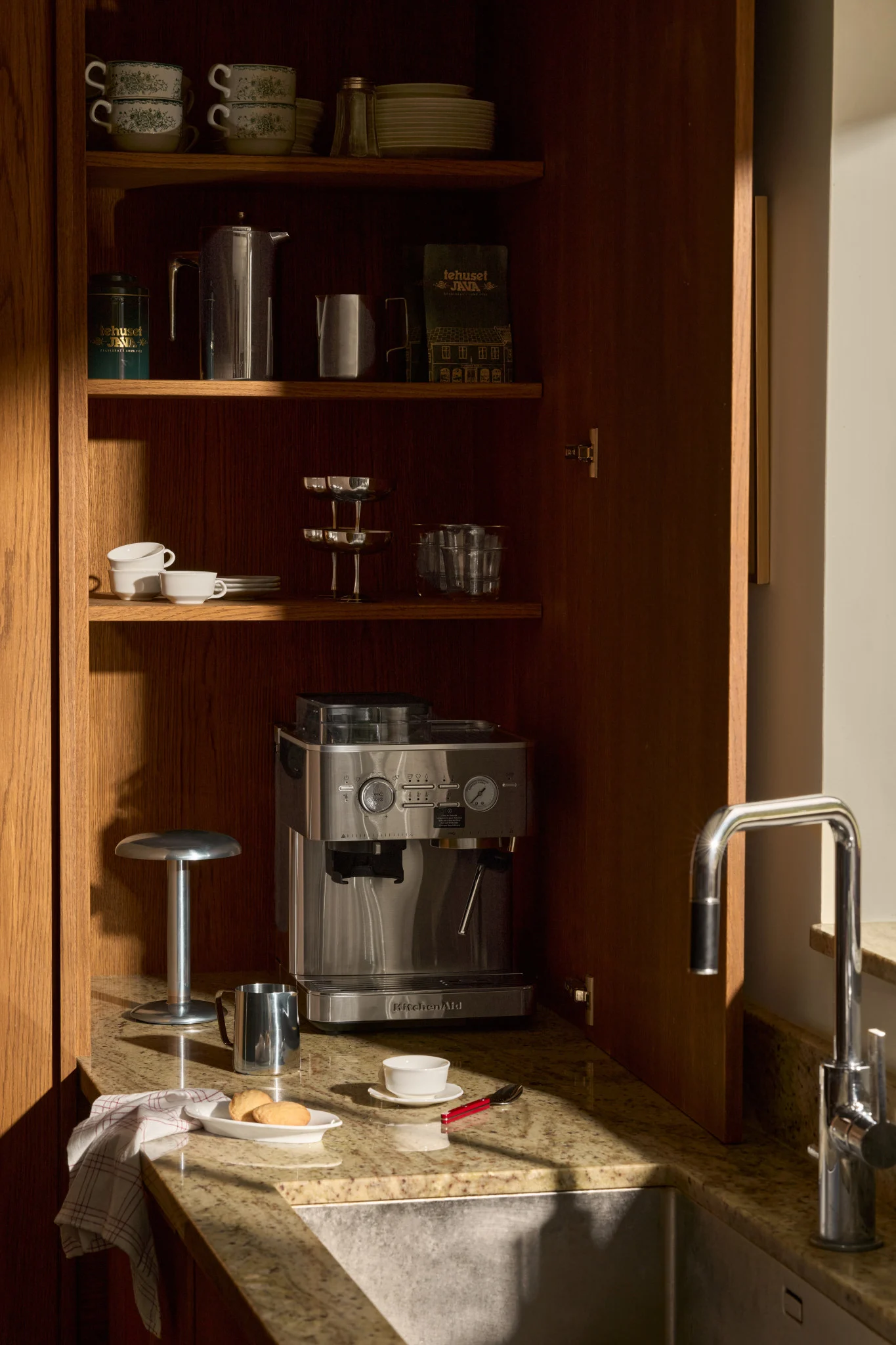 Ein offener Küchenschrank aus Holz mit Espressomaschine, Kaffeezubehör, gestapelten Tassen und Gläsern auf Regalen neben einem Waschbecken.