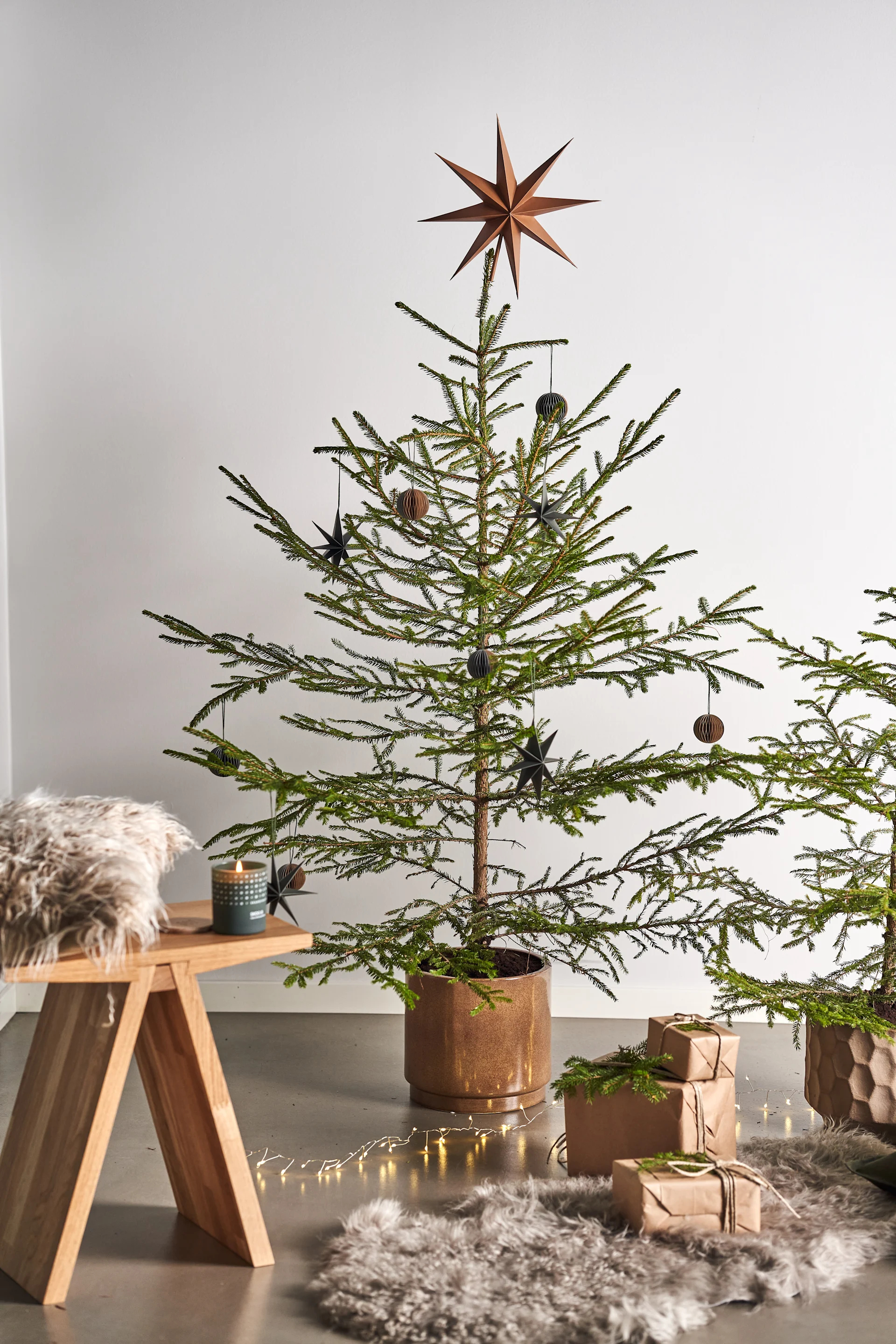 Dekorieren Sie Ihren Baum dieses Jahr mit dem Weihnachtsbaumschmuck 2021 in den 4 verschiedenen Stilen der Nest Trends – Nurture, Share, Boost und Cultivate. Hier sehen Sie eine Sakndinavisk Duftkerze mit dem Geruch des Waldes auf dem Angle Hocker von Muubs, welcher neben auf einem Schaffellteppich liegenden Geschenken steht. 