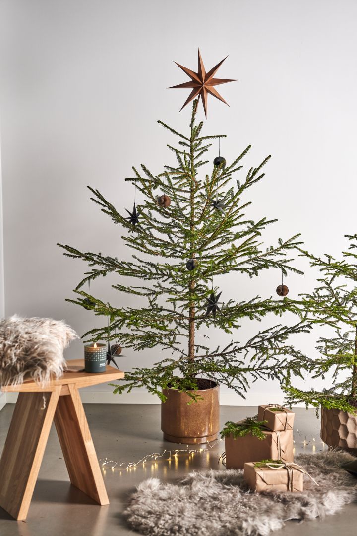 Dekorieren Sie Ihren Baum dieses Jahr mit dem Weihnachtsbaumschmuck 2021 in den 4 verschiedenen Stilen der Nest Trends – Nurture, Share, Boost und Cultivate. Hier sehen Sie eine Sakndinavisk Duftkerze mit dem Geruch des Waldes auf dem Angle Hocker von Muubs, welcher neben auf einem Schaffellteppich liegenden Geschenken steht. 