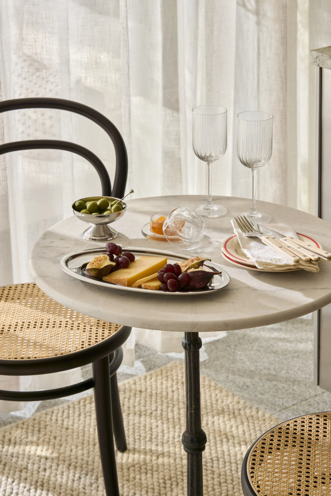 Ein Marmortisch mit Käse, Feigen, Trauben, Oliven, Marmelade, Gläsern und Besteck, mit Korbstühlen.