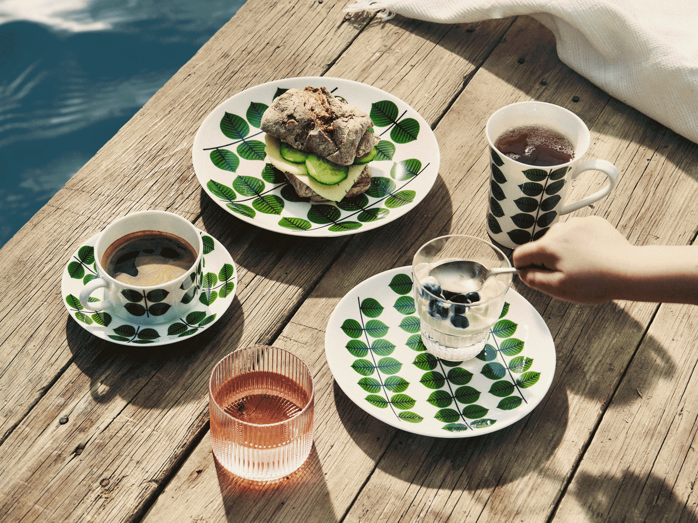 Frühstück auf Holzsteg: Geschirr mit Blattmuster, Sandwich, Kaffee, Tee, Joghurt mit Blaubeeren und pinkes Getränk.
