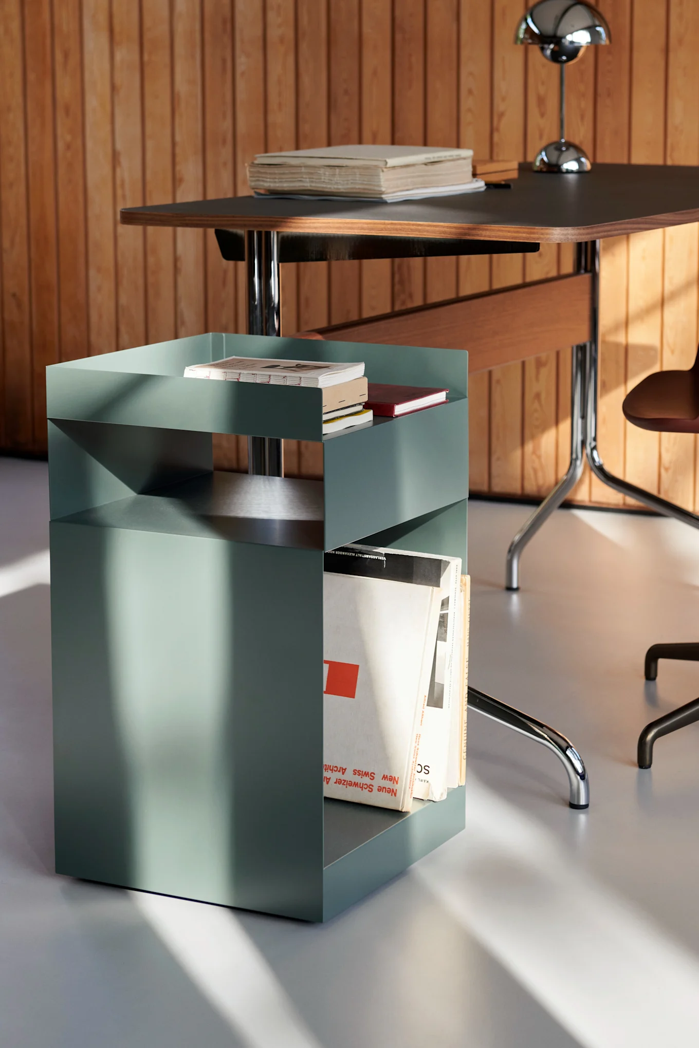 Salbeigrüner Metall-Beistelltisch mit geometrischem Design, mit Büchern und Zeitschriften, neben einem Holzschreibtisch mit einer Silberlampe.