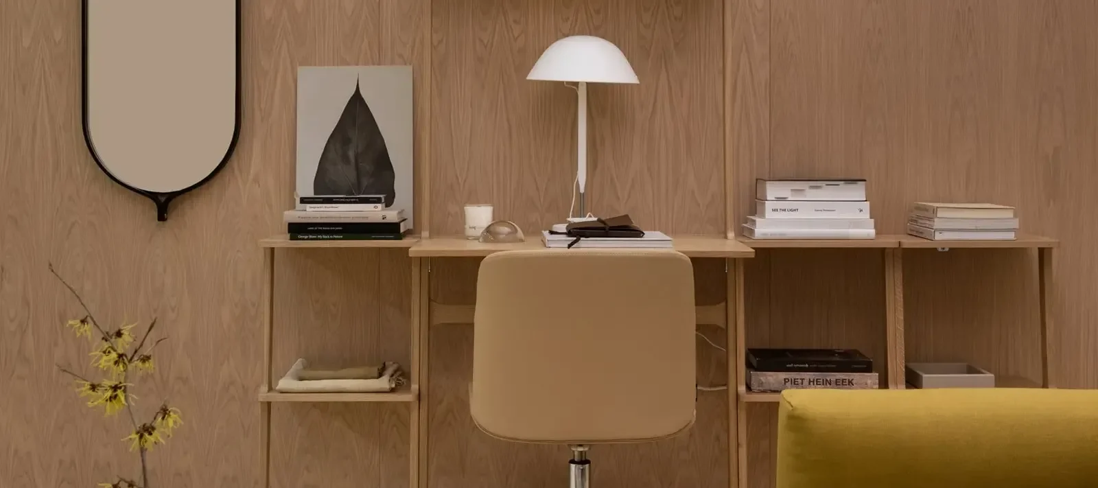 Arbeitsplatz mit hellem Holzschreibtisch, beigem Stuhl, weißer Schreibtischlampe, Büchern, Spiegel und einer Pflanze mit gelben Blüten.