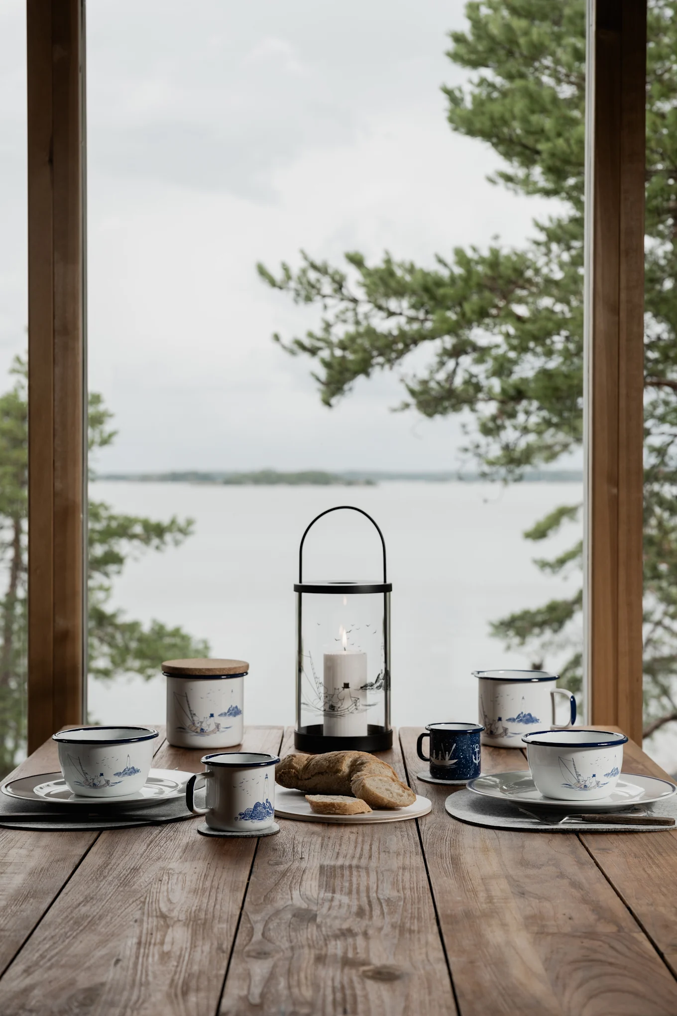 Windlicht für den Tisch: Ein Holztisch vor einem schwedischen See, gedeckt mit Kaffeetassen und dem Mumin Windlicht von Muurla.