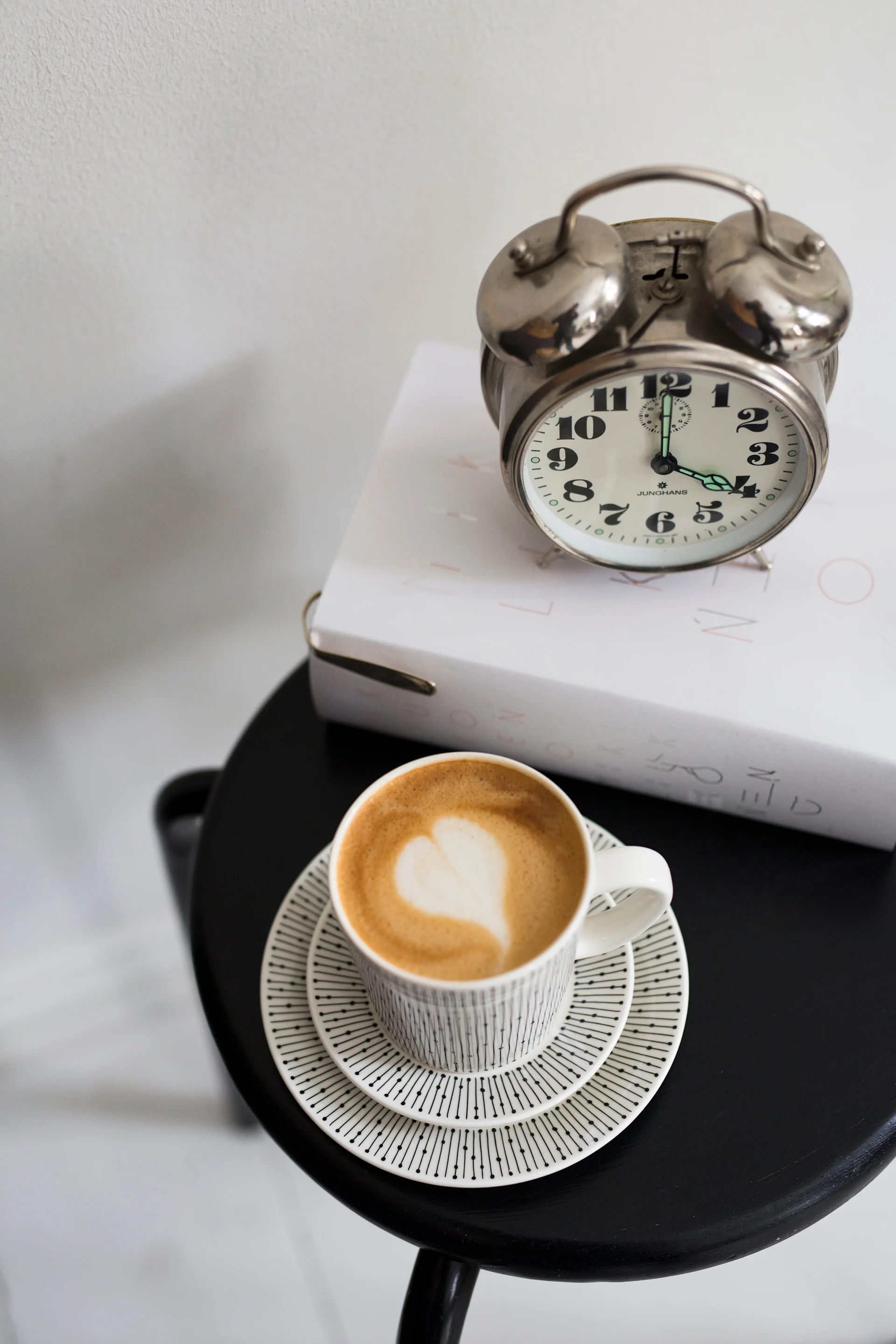 Das Bild zeigt einen Hocker, auf dem ein Buch liegt, darauf ein Wecker. Davor steht die Mainio Sarastus Tasse von Arabia mit Kaffee und herzförmigem Schaum - genau richtig für eine entspannte Fika.