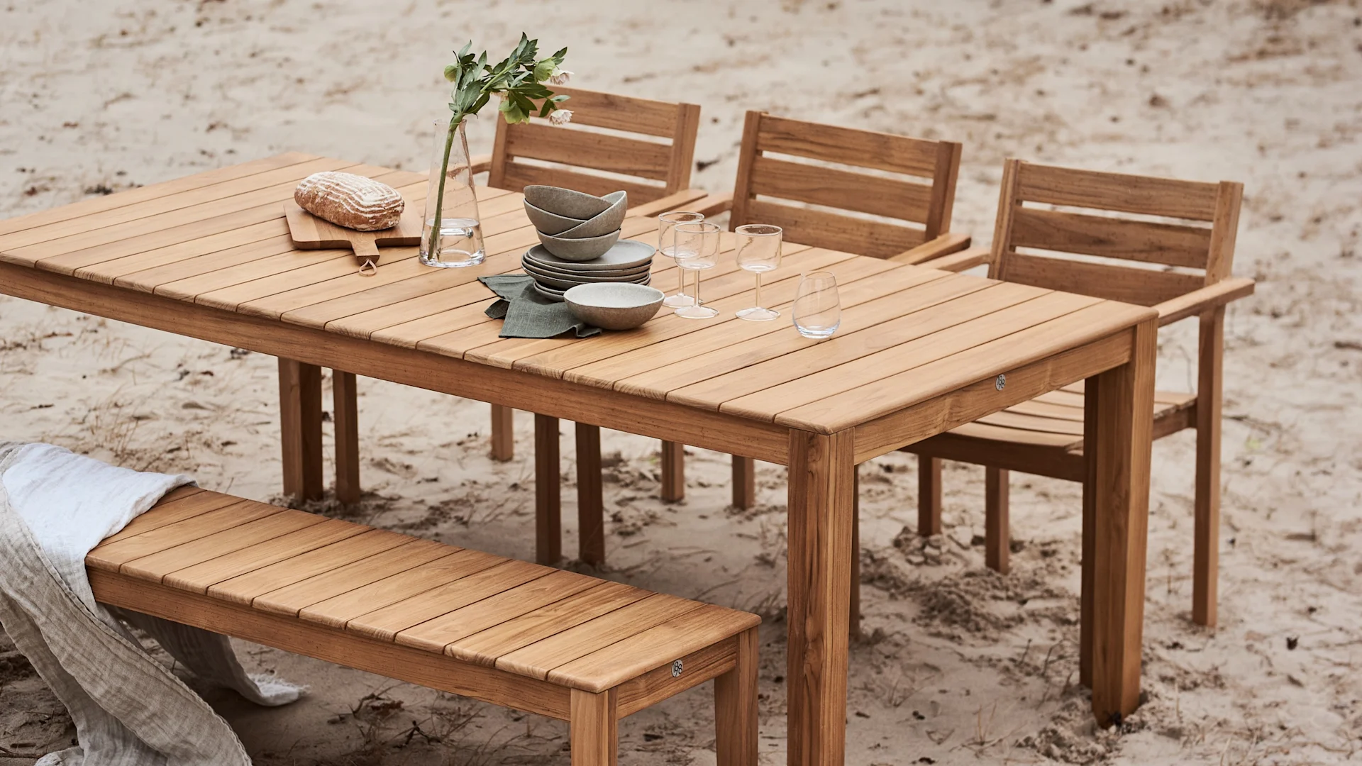 Hölzerner Gartentisch gedeckt mit Geschirr, Gläsern, einer Blumenvase und Brot, umgeben von Stühlen und einer Bank im Sand.