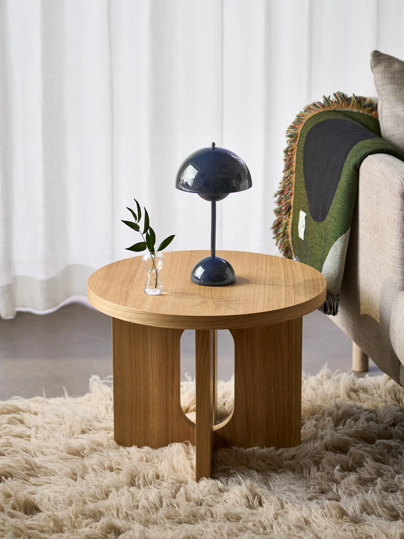 Runder Holznebentisch mit dunkelblauer Pilzlampe und kleiner Pflanze, neben einem Sessel mit grüner Decke, auf einem flauschigen Teppich.