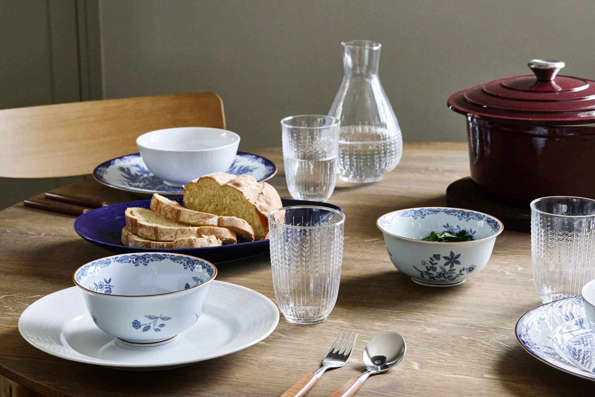 Ein Holztisch gedeckt mit blau-weiß gemusterten Schüsseln, Tellern, geschnittenem Brot, Wassergläsern und einem roten Topf mit Deckel.