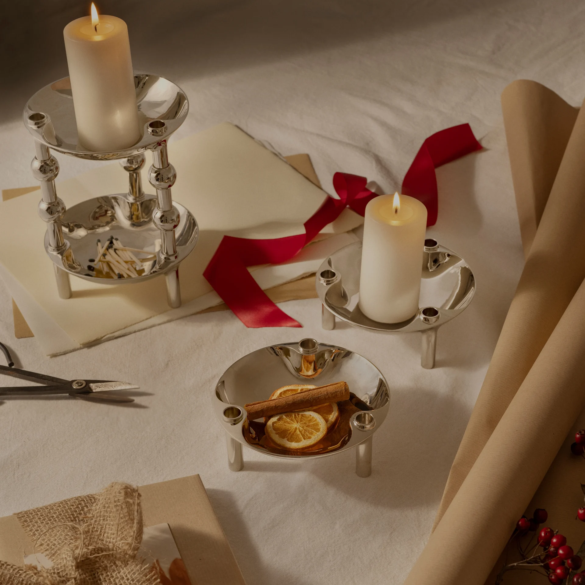 Drei silberne Kerzenhalter, zwei mit brennenden Stumpenkerzen, einer mit getrockneten Orangen und Zimt, arrangiert mit Geschenkpapier und Band.