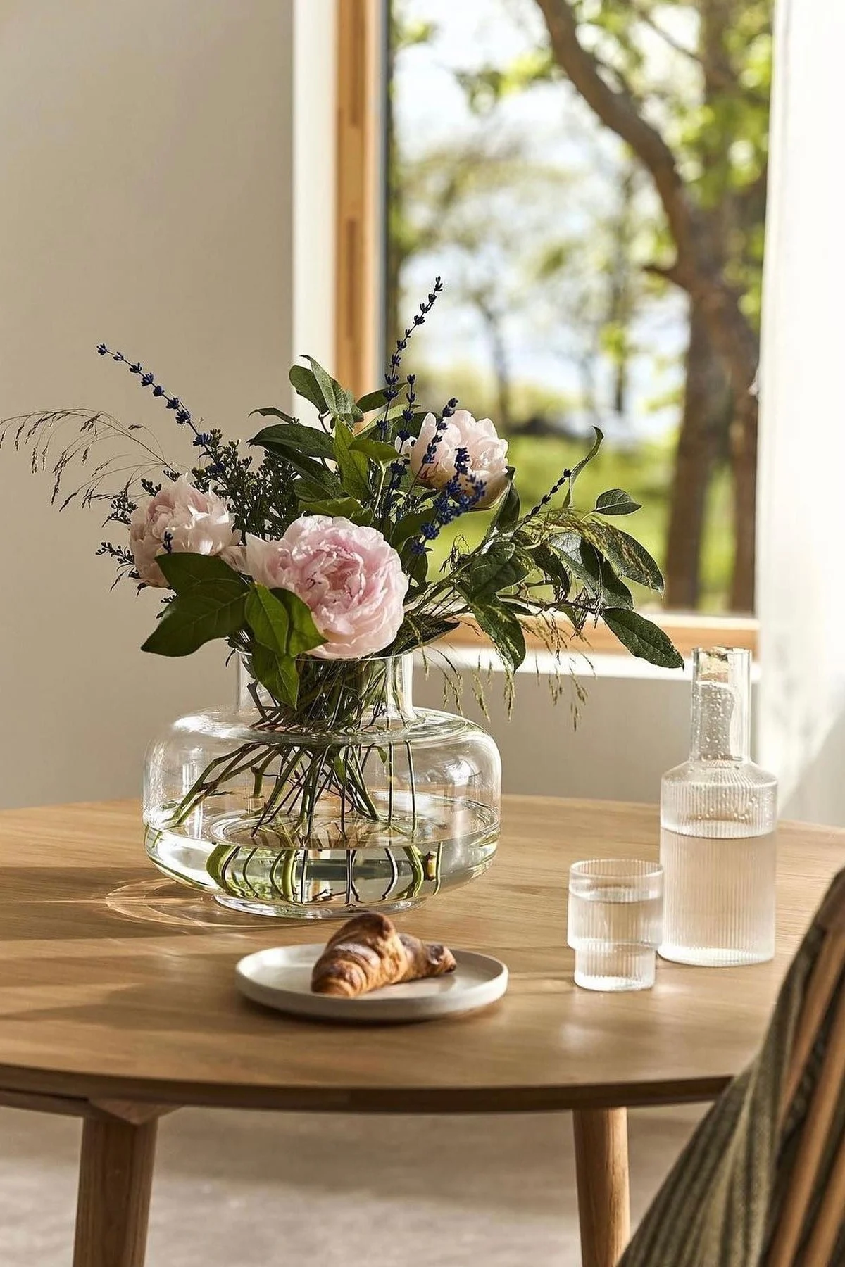 Auf einem mit Karaffe und Croissant gedeckten Tisch steht die Urna Vase von Marimekko mit Pfingstrosen.