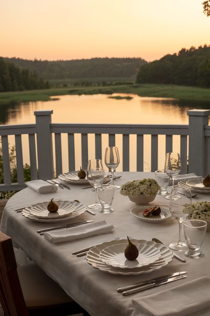 Ein Tisch bei Sonnenuntergang auf einer Terrasse mit Blick auf einen See. Der Tisch ist in Weiß mit Tellern aus der Oyster-Kollektion von Mateus gedeckt.