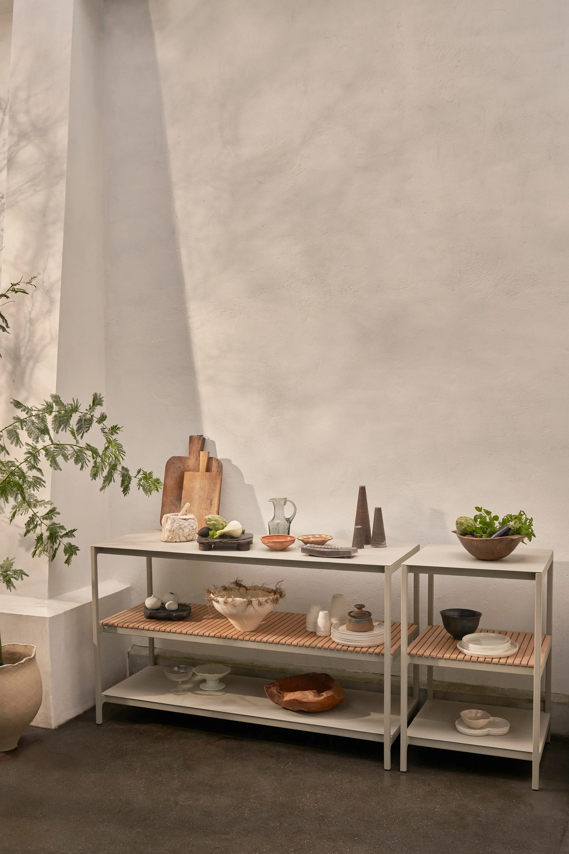 Zwei moderne graue Regale mit Holzlattenauflagen präsentieren diverse Küchenutensilien, Keramikschalen, frisches Gemüse und Dekoartikel vor einer hellen Wand.