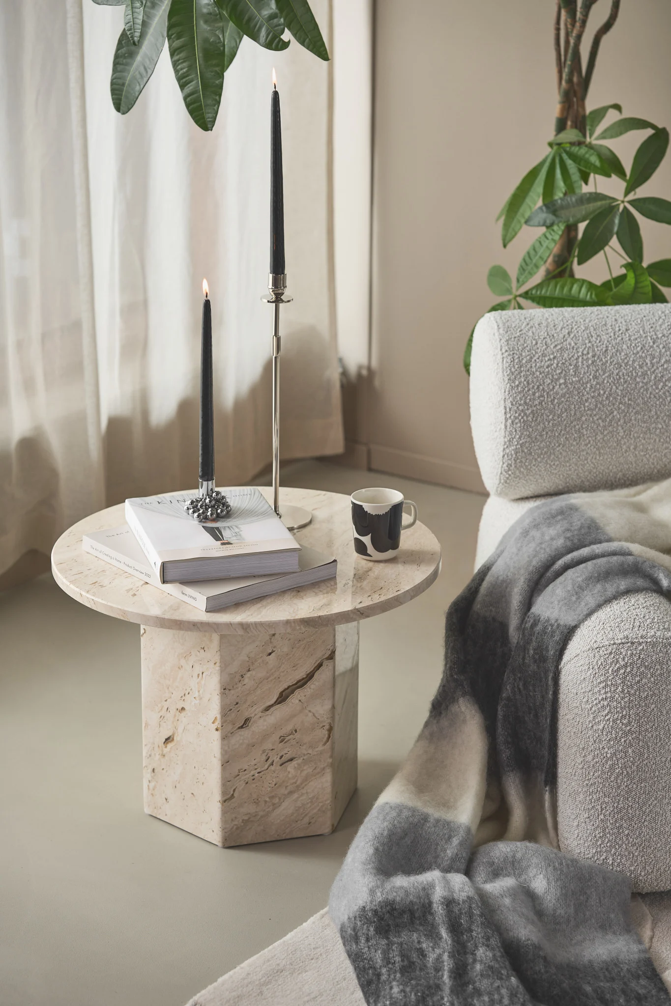 Gemütliches Zimmer mit Marmor-Beistelltisch, zwei brennenden schwarzen Kerzen, Büchern und einer Tasse neben einem Sessel mit Decke.