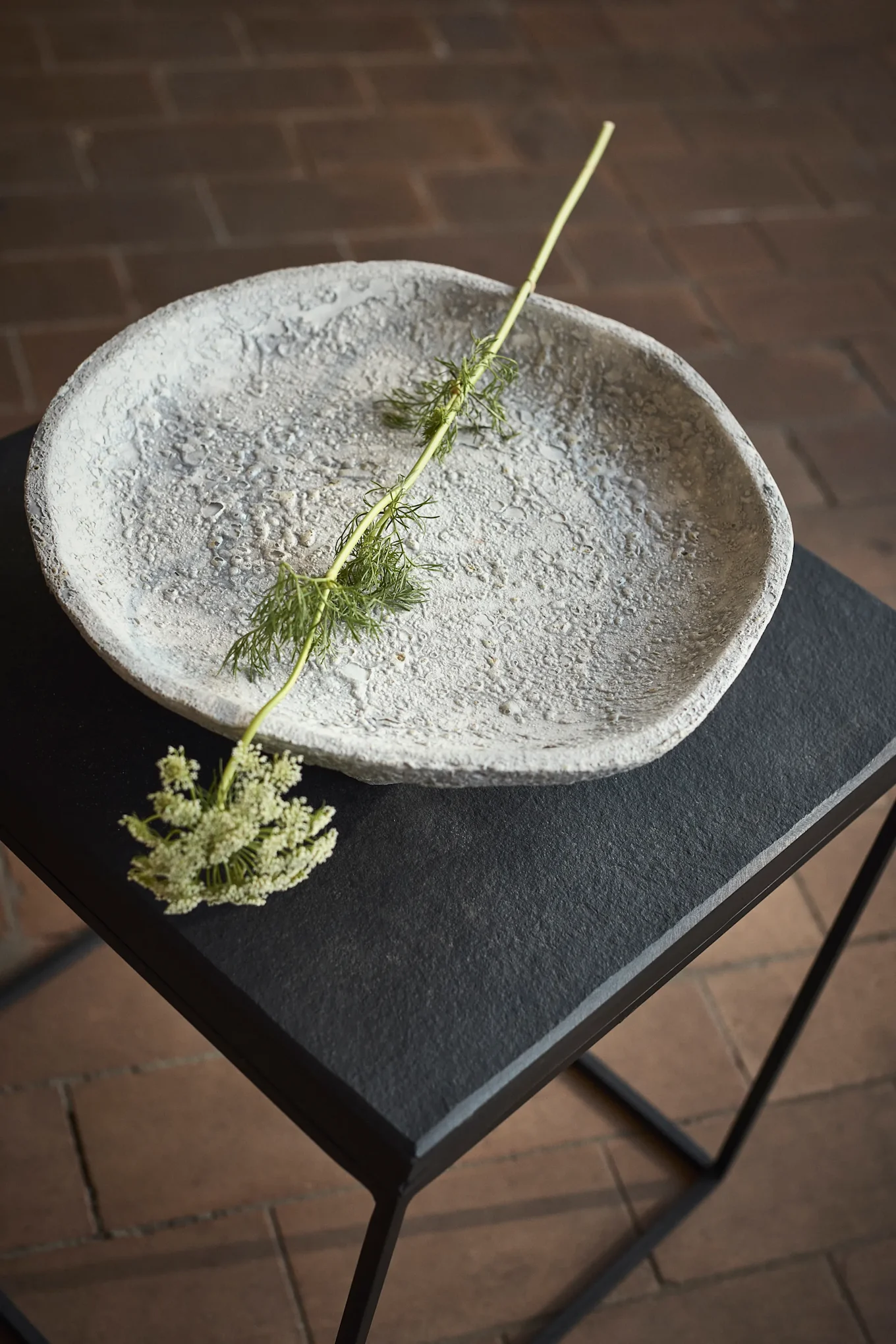 Strukturierte graue Schale auf einem schwarzen Tisch mit grünem Stiel und kleinen weißen Blüten.
