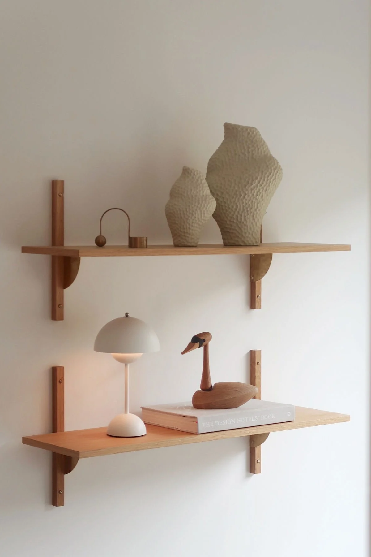 Zwei helle Wandregale aus Holz mit Dekoration. Das obere Regal beherbergt zwei texturierte Vasen und Metallobjekte. Das untere Regal hat eine weiße Lampe, einen Holzschwan und ein Buch.