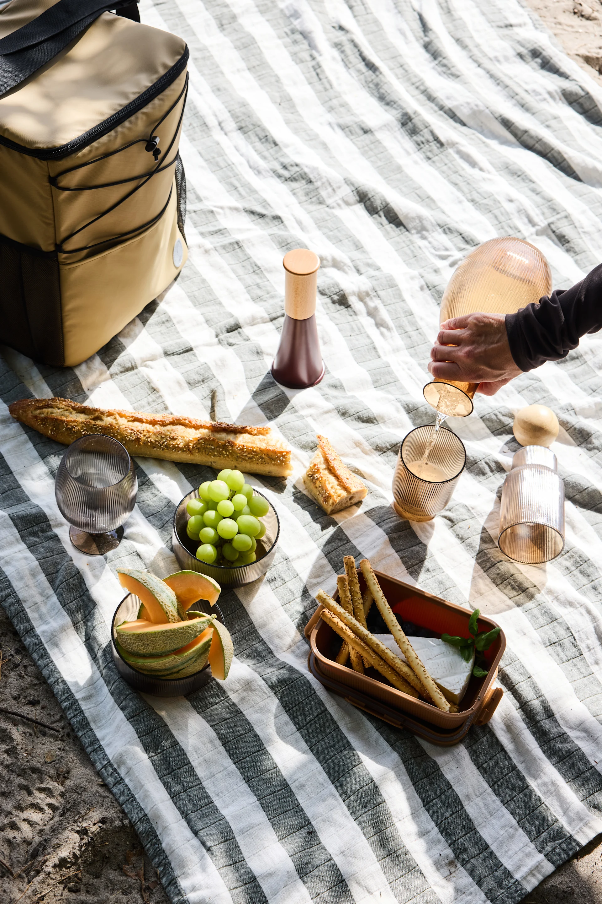 Blick von oben auf ein Picknick auf einer gestreiften Decke, mit einer Person, die aus einer geriffelten Karaffe gießt, Baguette, Obst, Käse und Grissini.