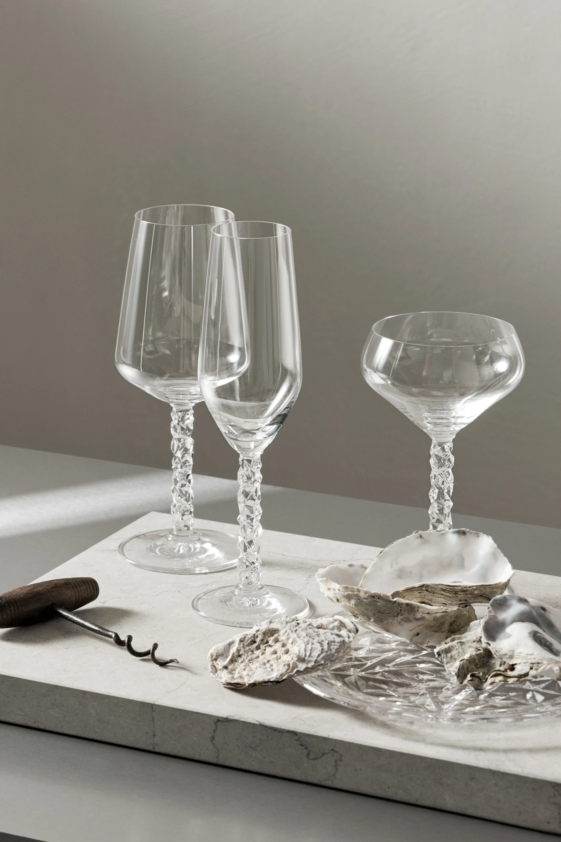 Drei klare Glasstielgläser mit facettierten Stielen, ein Korkenzieher und Austernschalen auf einer hellen Oberfläche.