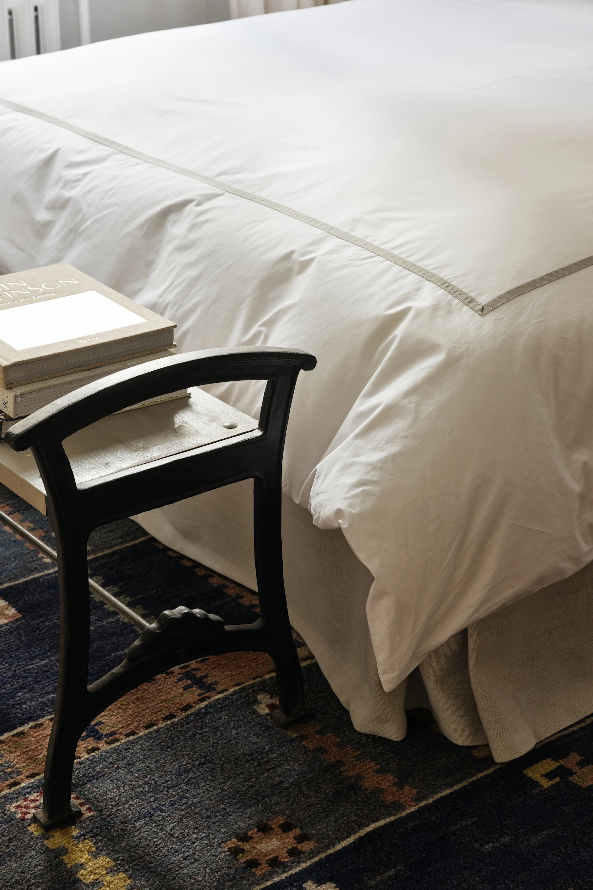 Ein Bett mit weißer Bettwäsche und einem grauen Streifen neben einem dunklen Hocker mit Büchern auf einem gemusterten blauen Teppich.