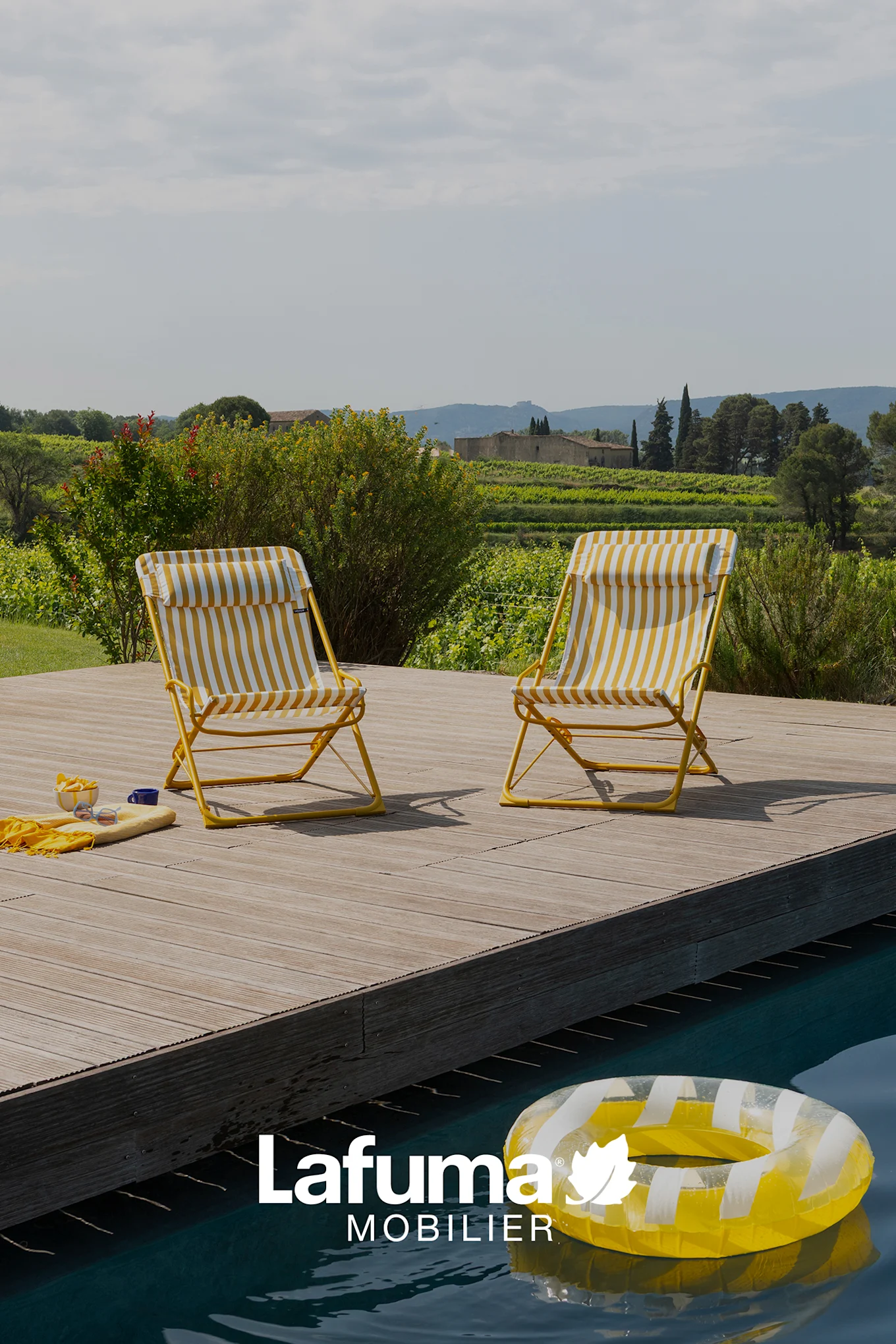 Zwei gelb-weiß gestreifte Klappstühle an einem Pool, mit einem Weinberg und Gebäuden im Hintergrund.