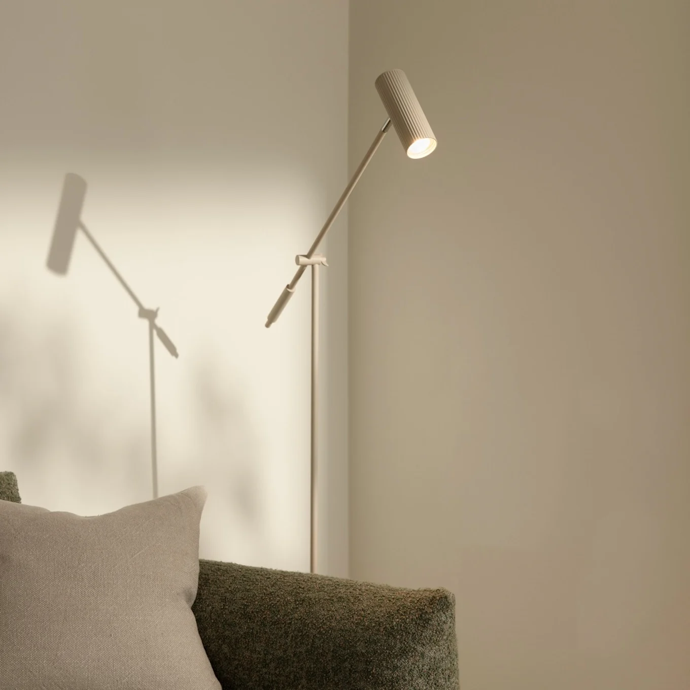 Eine moderne, schlanke Stehlampe mit geripptem Kopf, die eine helle Wand beleuchtet, neben einem grünen strukturierten Sofa mit beigem Kissen.