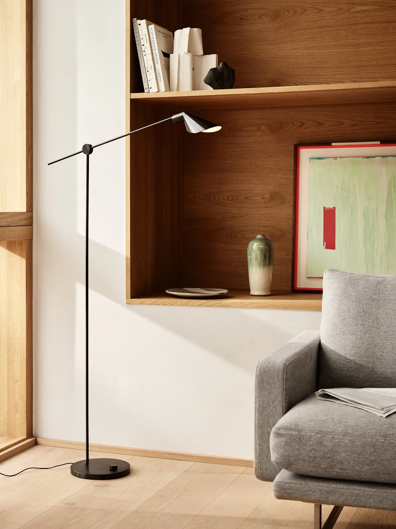 Ein modernes Wohnzimmer mit einer schlanken schwarzen Stehlampe, Holzregalen mit Büchern und Dekoration sowie einem Teil eines grauen Sofas.