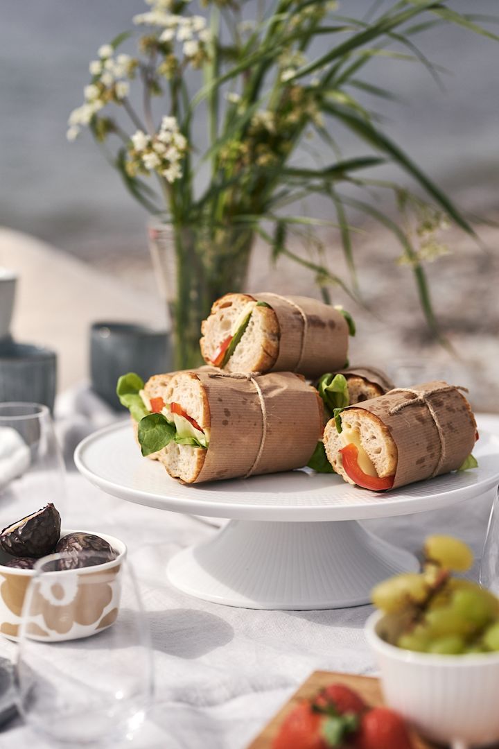 Picknick am Strand mit in Wachspapier eingewickelten Sandwiches auf einer weißen Kuchenplatte. 