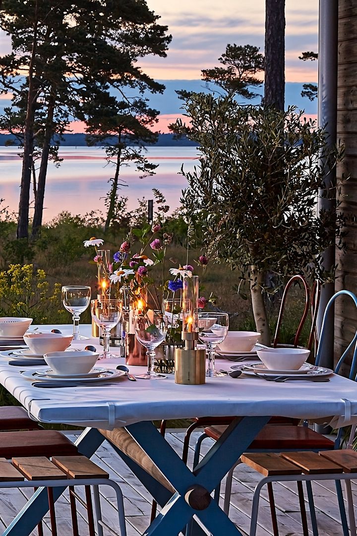 Laternen und Windlichter für draußen – hier sehen Sie einen Holztisch auf einer Terrasse mit dem Meer im Hintergrund während des Sonnenuntergangs. Der Tisch ist gedeckt und unter anderem mit der Patina Mini-Ölleuchte von Klong dekoriert.